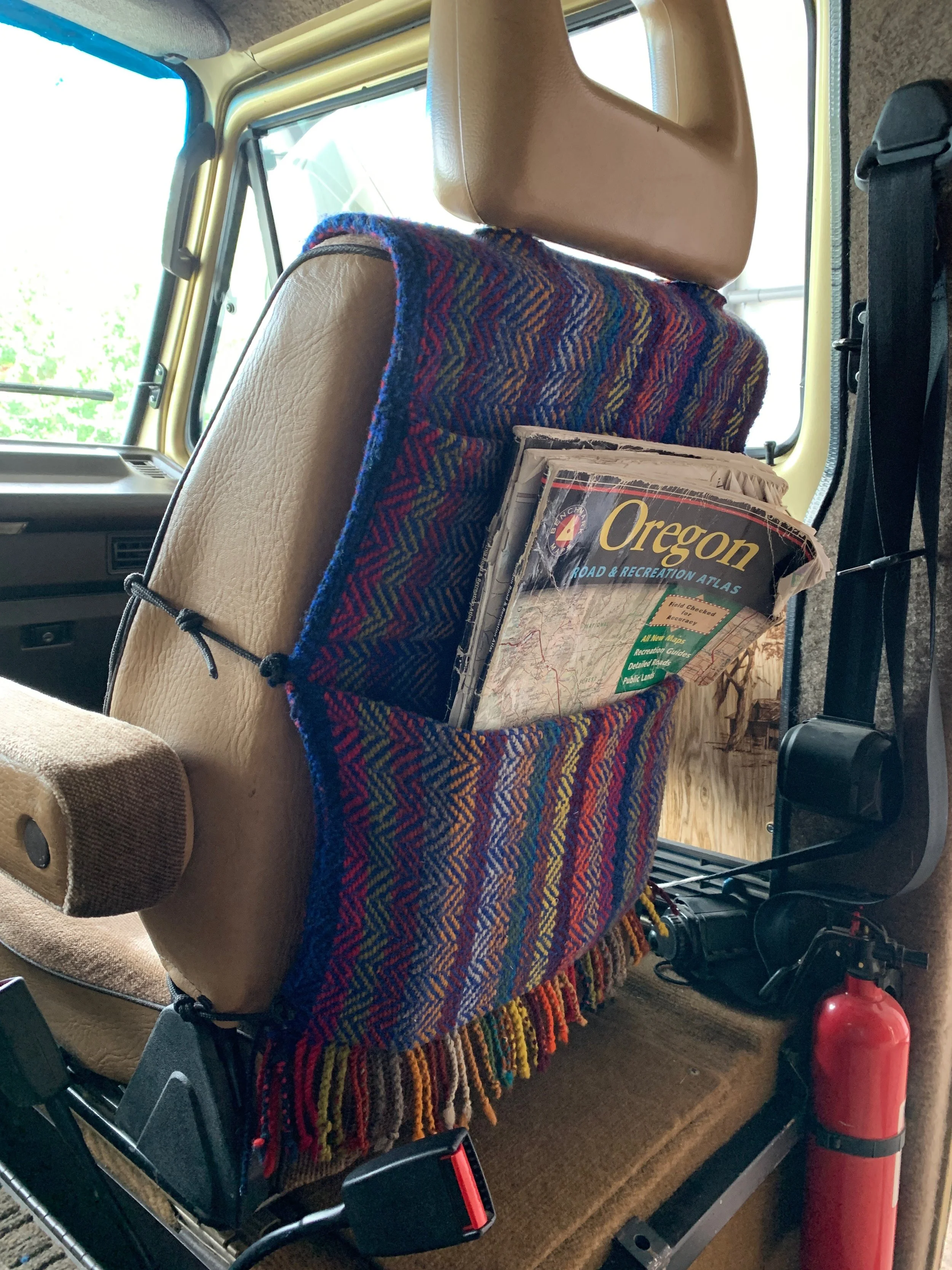 View inside a vehicle showing the back of a beige leather seat, a colorful woven blanket with fringes, a folded newspaper titled Oregon Road & Recreation Atlas in a mesh pocket, and a red fire extinguisher on the floor near the seat.