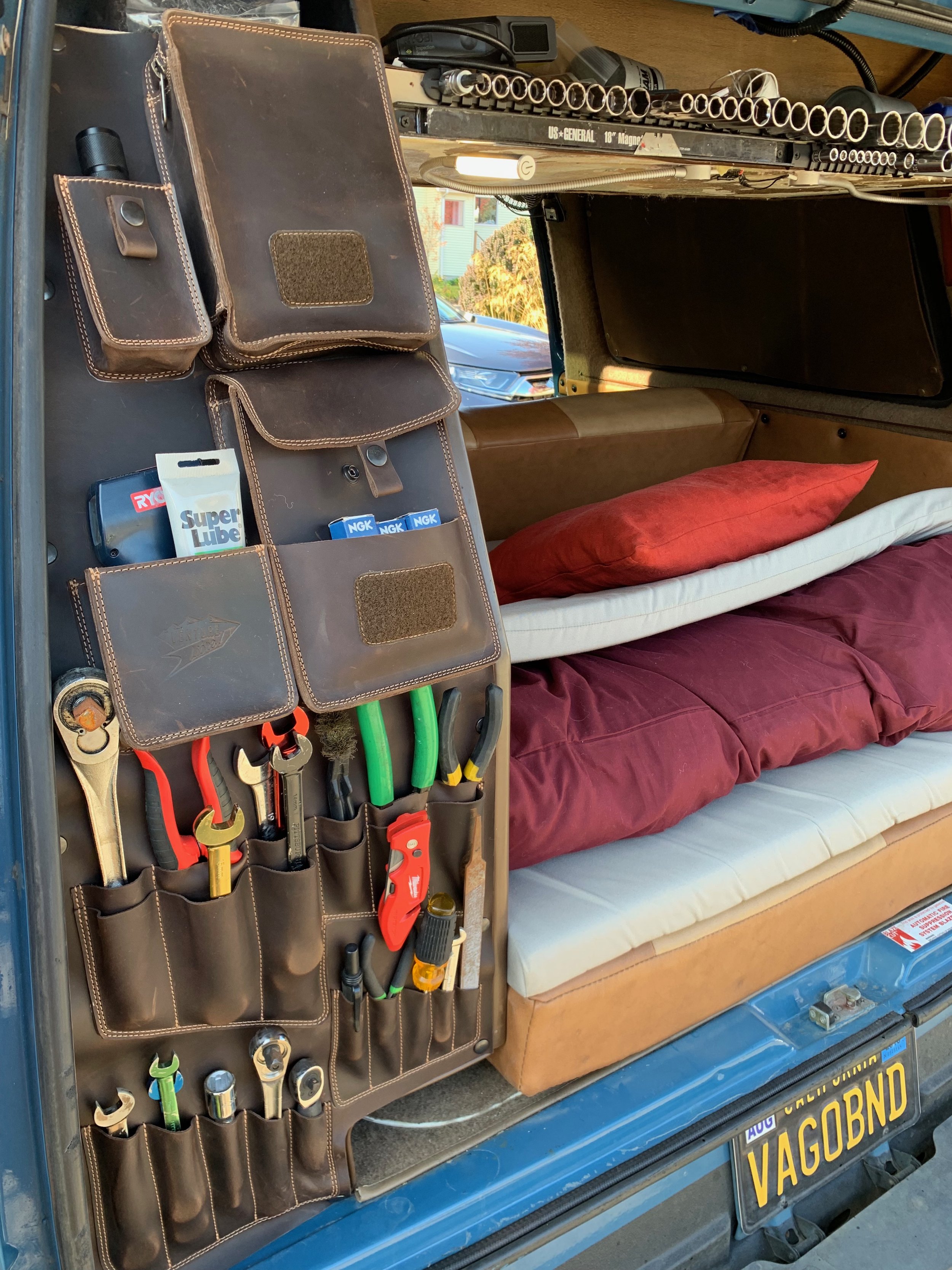 The image shows the open back of a vehicle with a tool organizer and various tools and supplies, including wrenches, screwdrivers, and oil. There is a sofa with pillows inside the vehicle, and a custom license plate reading 'VAGOBND' is visible at th