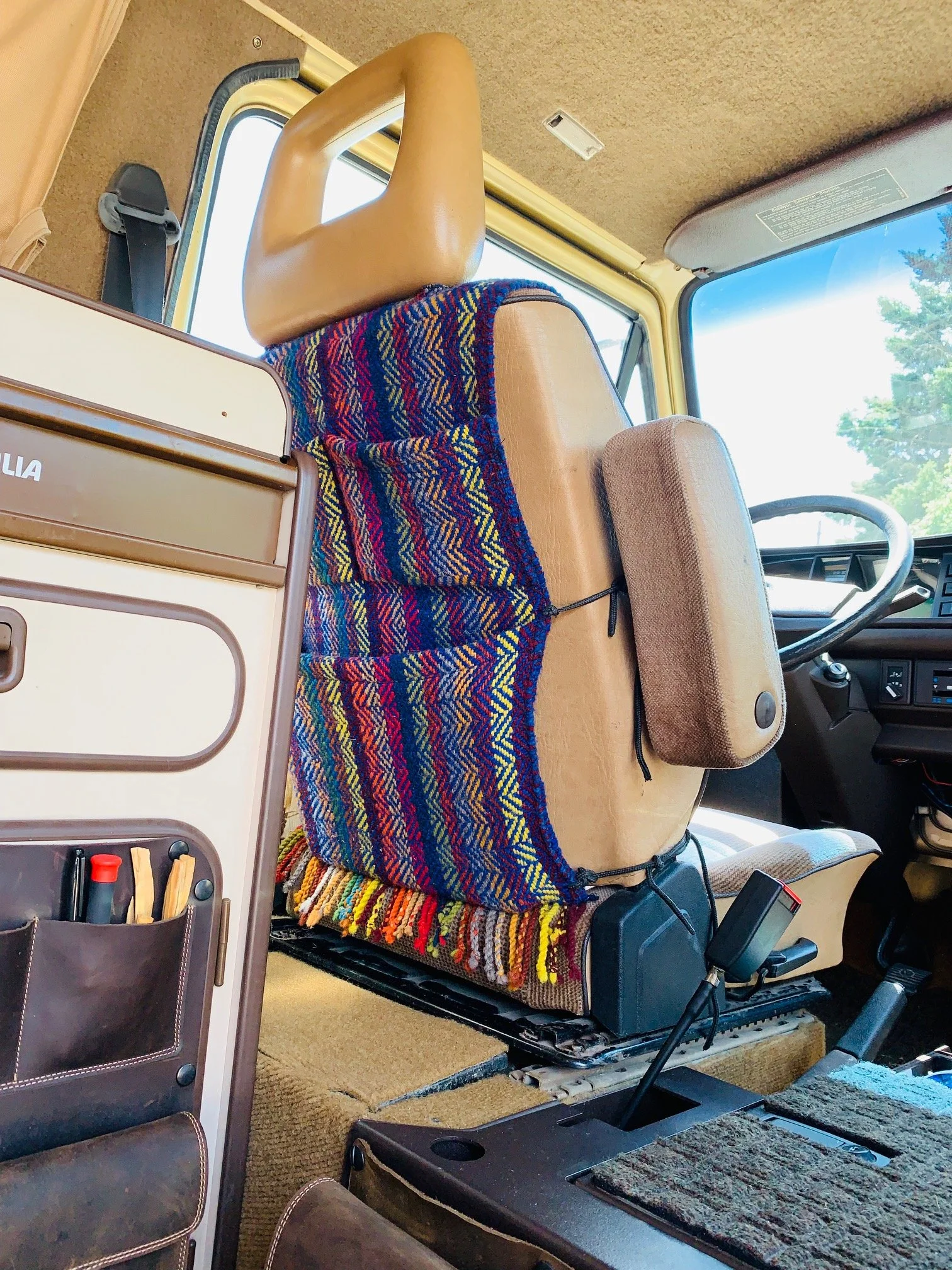 Inside the cab of a bus, featuring the driver's seat and dashboard, with part of a passenger seat visible, and a colorful woven blanket draped over the back of the seat.