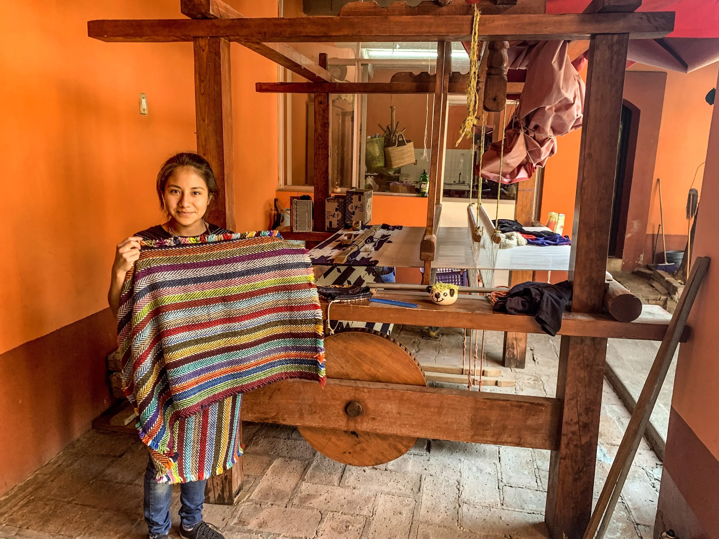 Young girl with dark hair holding colorful woven textile, standing inside a room with orange-painted wall and a large wooden loom.