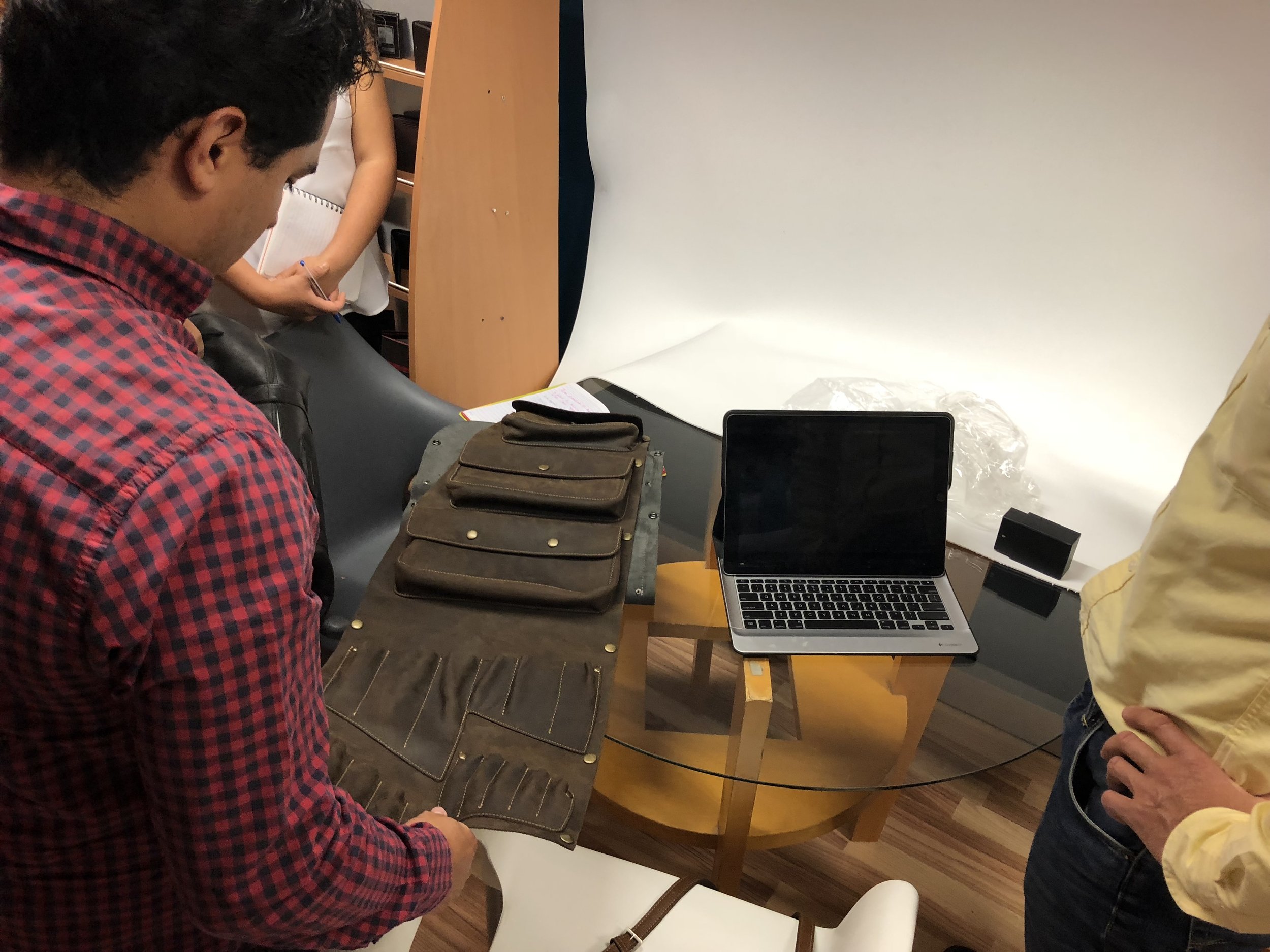 People gathered around a table with a brown leather bag, a laptop, and notebooks, engaging in a discussion or meeting indoors.