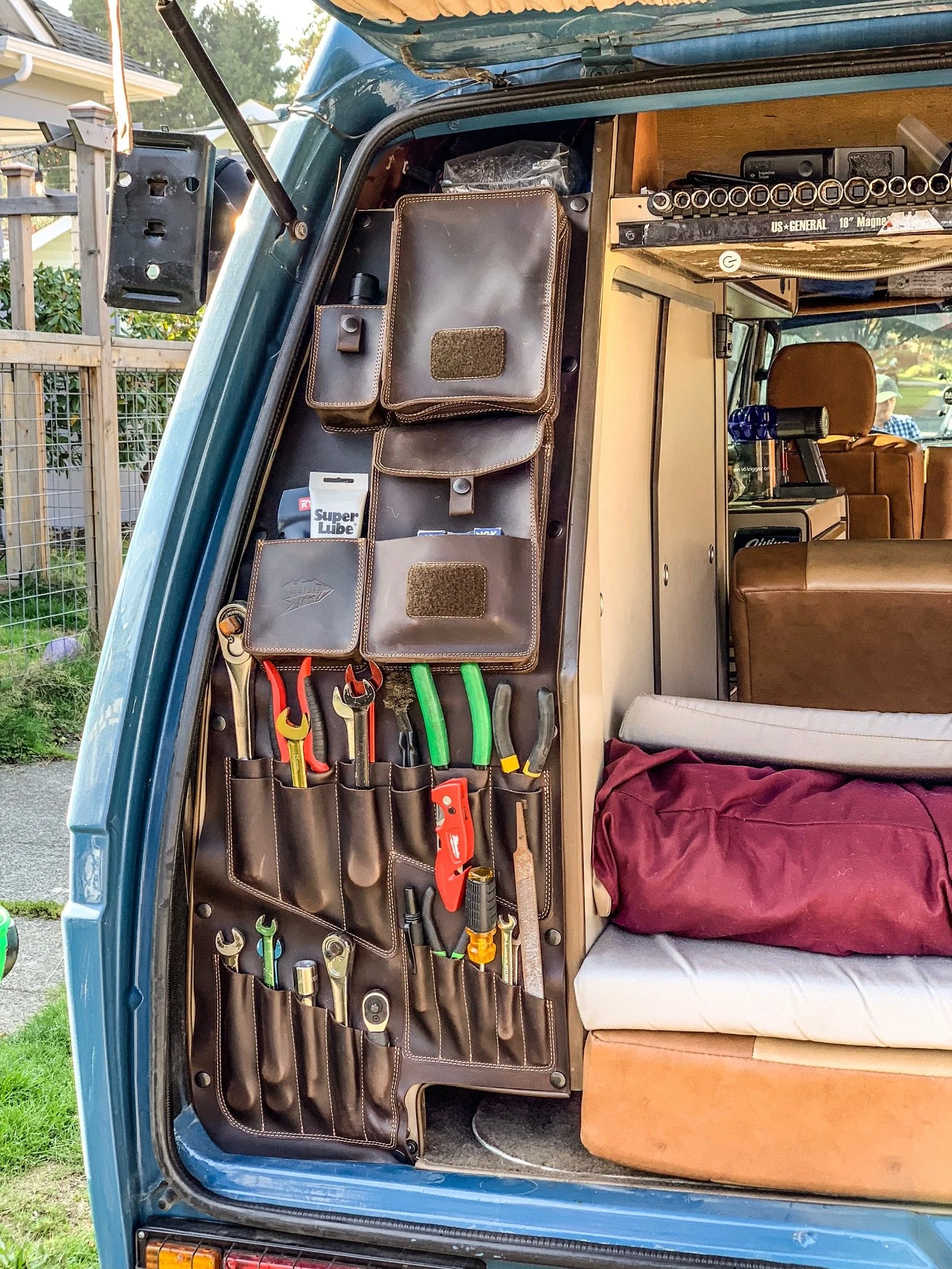 Inside the rear of a blue van, organized with a leather tool roll containing various hand tools, a cardboard box, and a belt pouch, with a water bottle and a brown couch visible in the background.