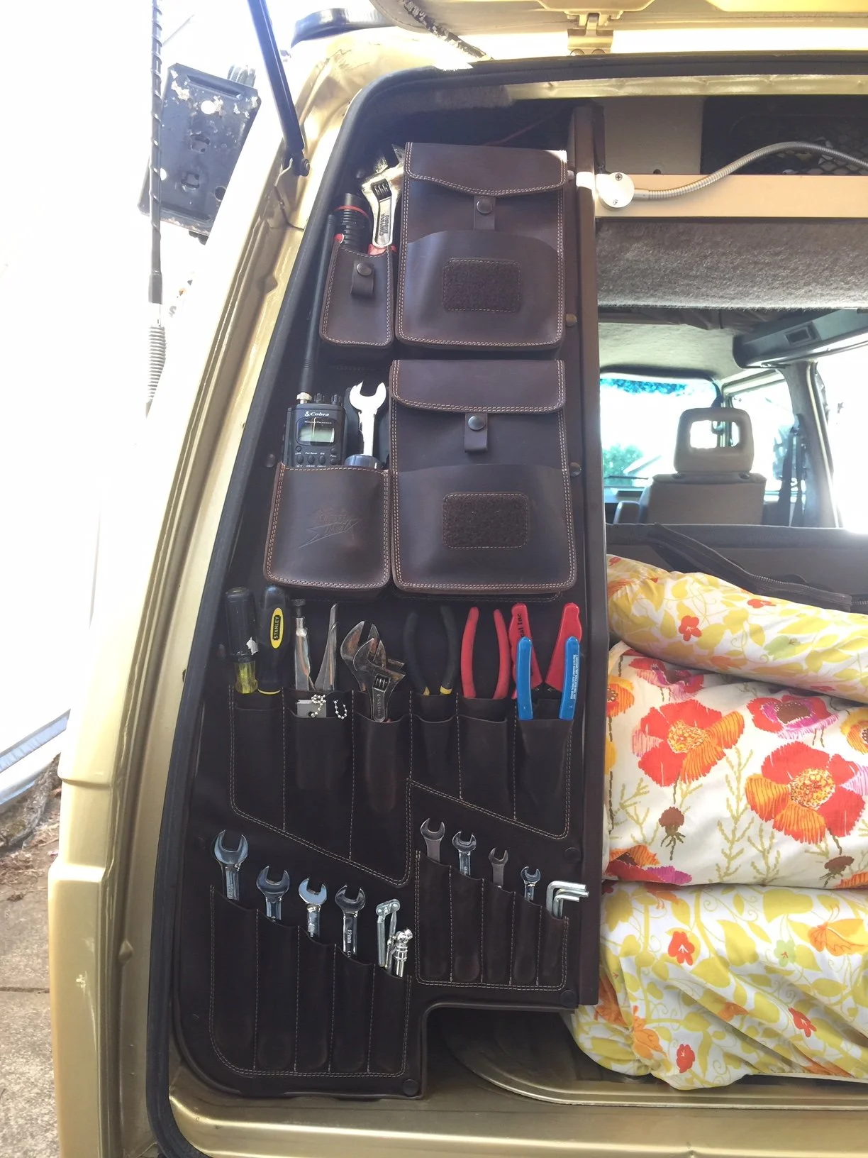 Organizer with tools hanging on the side of a vehicle, including wrenches, screwdrivers, pliers, and a tape measure, with a bed covered in a floral blanket nearby inside the vehicle.
