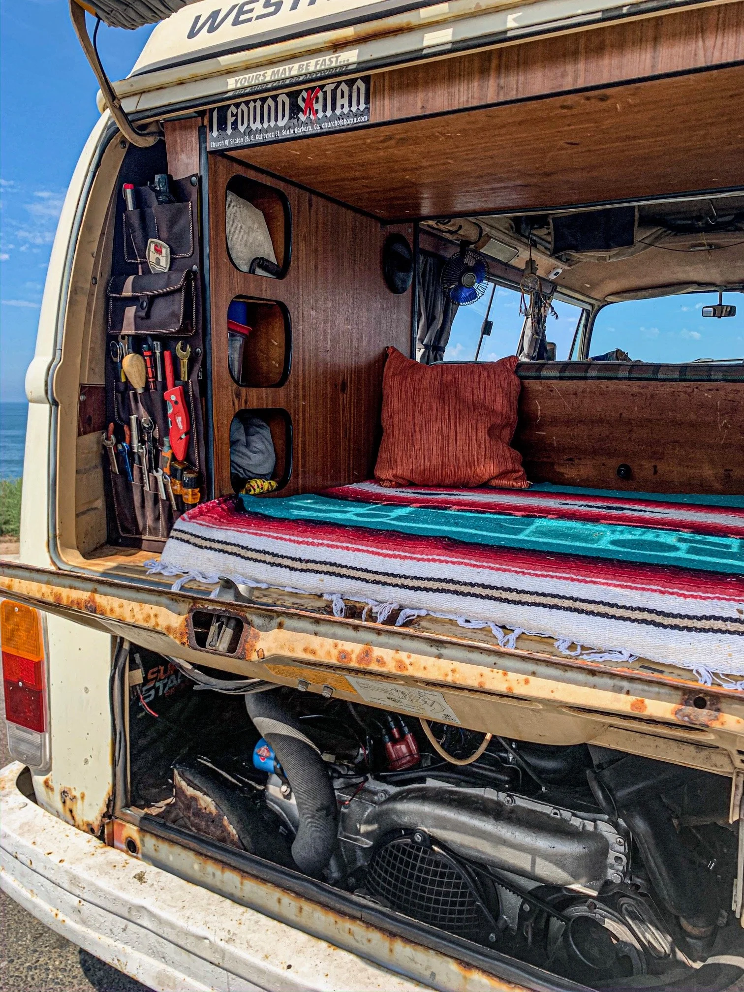 The image shows the back of a rusted camper van with its rear door open, revealing a wooden interior with tools and a colorful striped blanket, and a motorcycle stored underneath.