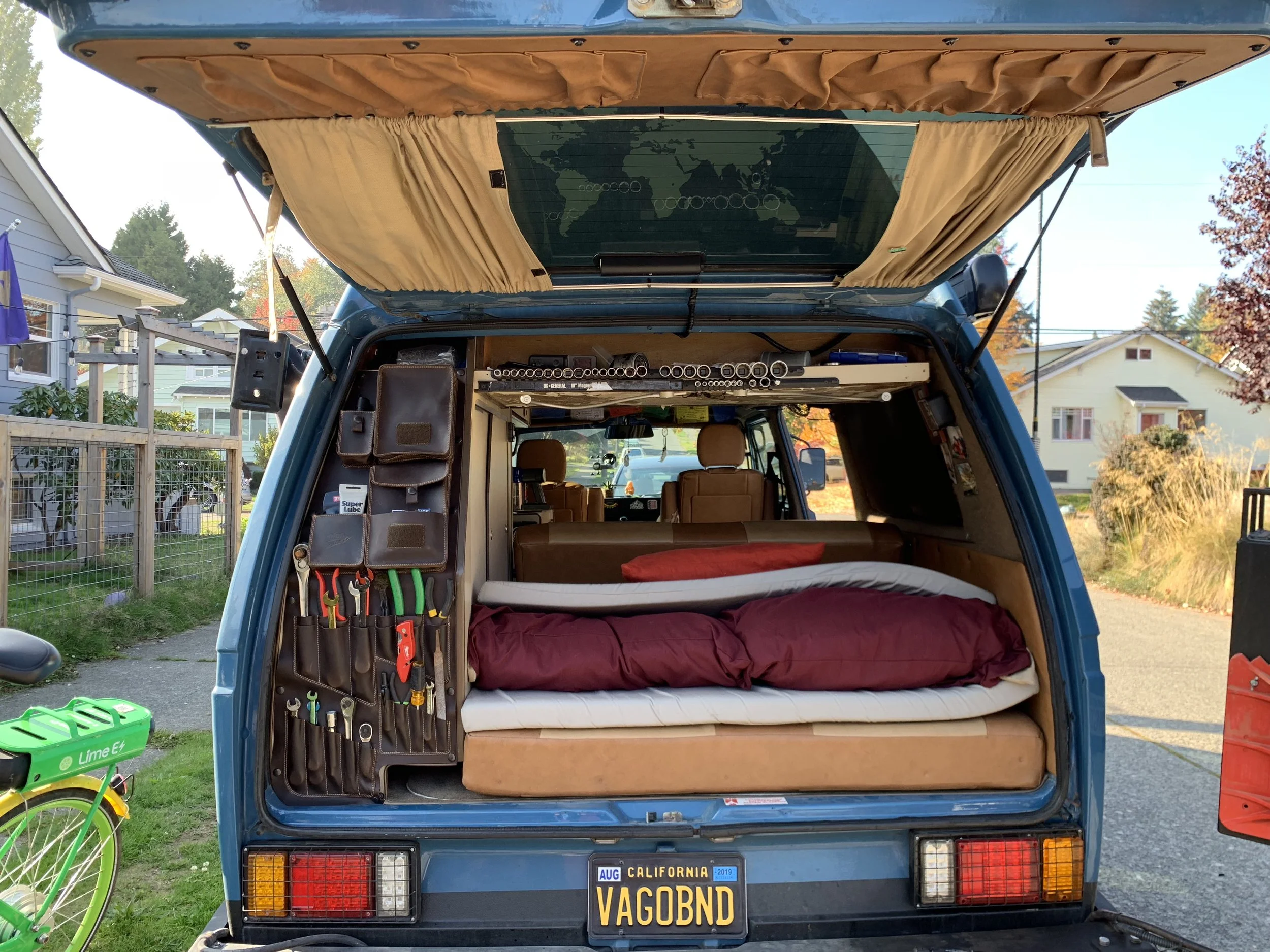Back of blue van with open hatch revealing the interior with a bed, tool organizer, and wooden shelf with socket wrenches. California license plate reads 'VAGOBND'.