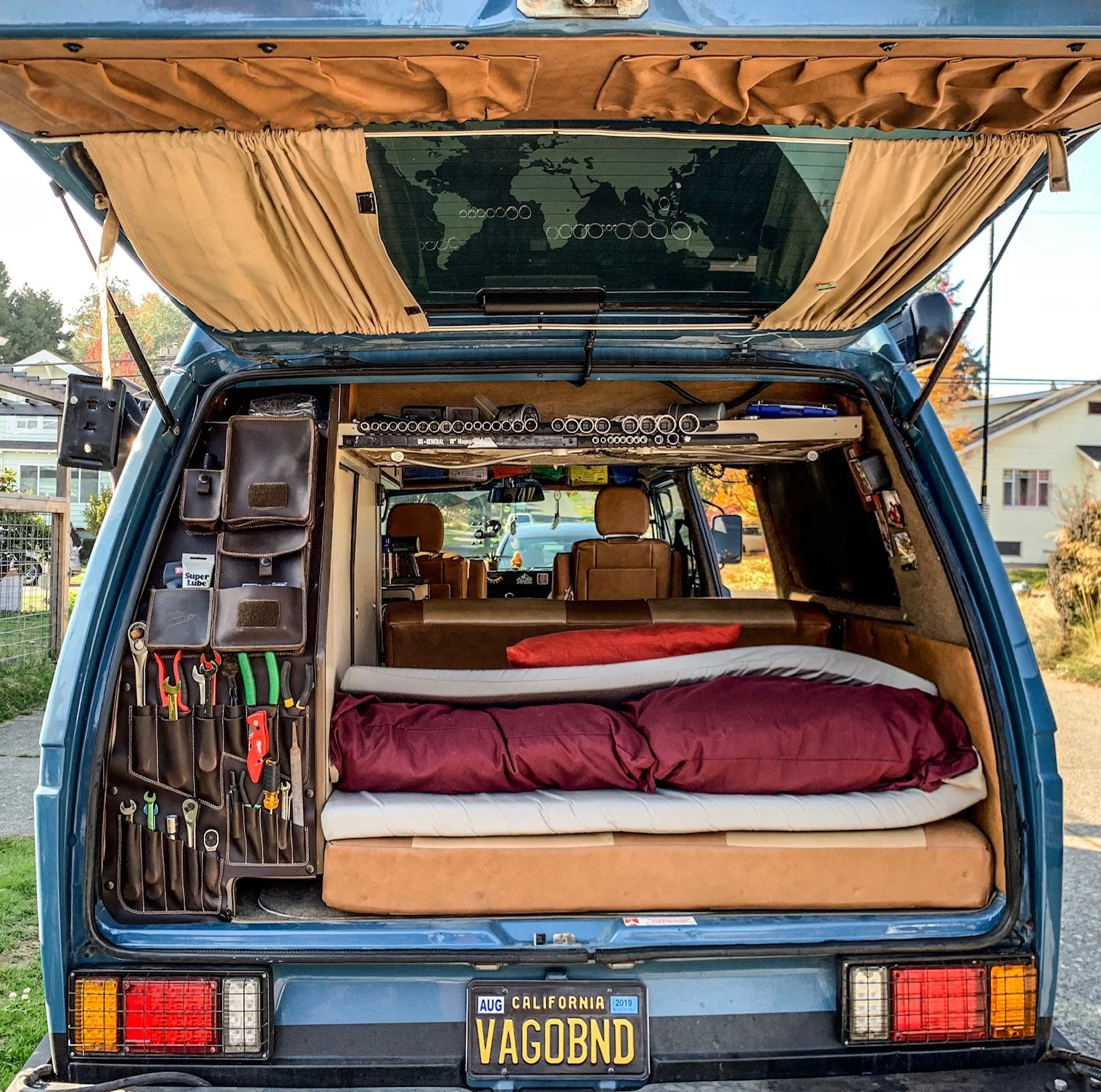 The rear of a blue camper van with an open hatchback showing interior storage and a bed. The interior includes organized tool pouches on the left, a bed with red bedding, and a view of the front seats and dashboard. The back of the van has a Californ