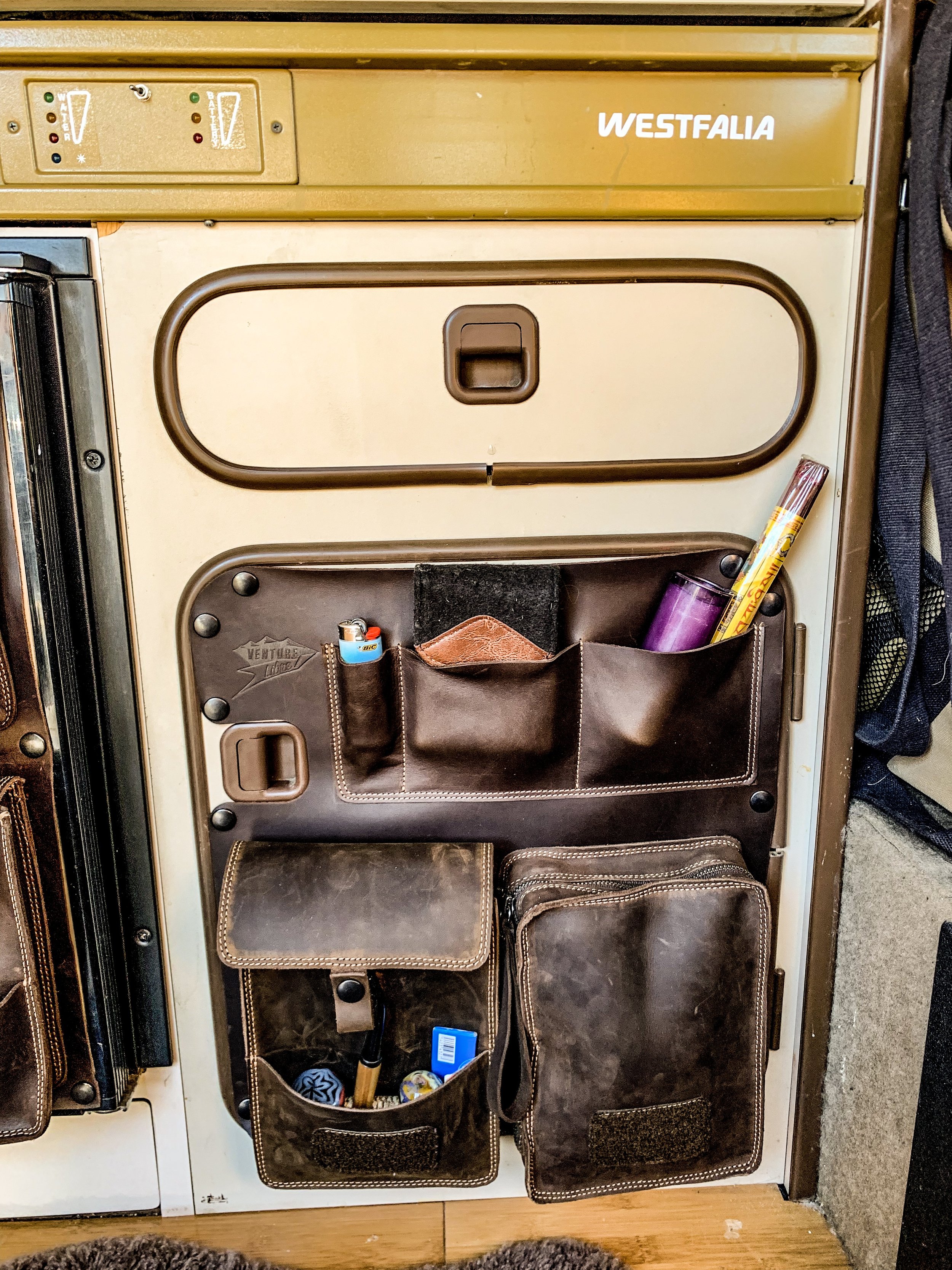The image shows a vintage Westfalia camper van refrigerator with a storage pocket attached to the door, holding small items like a purple cup, a lighter, a small container, and a yellow item. The refrigerator has thermometer gauges on top, and the ov
