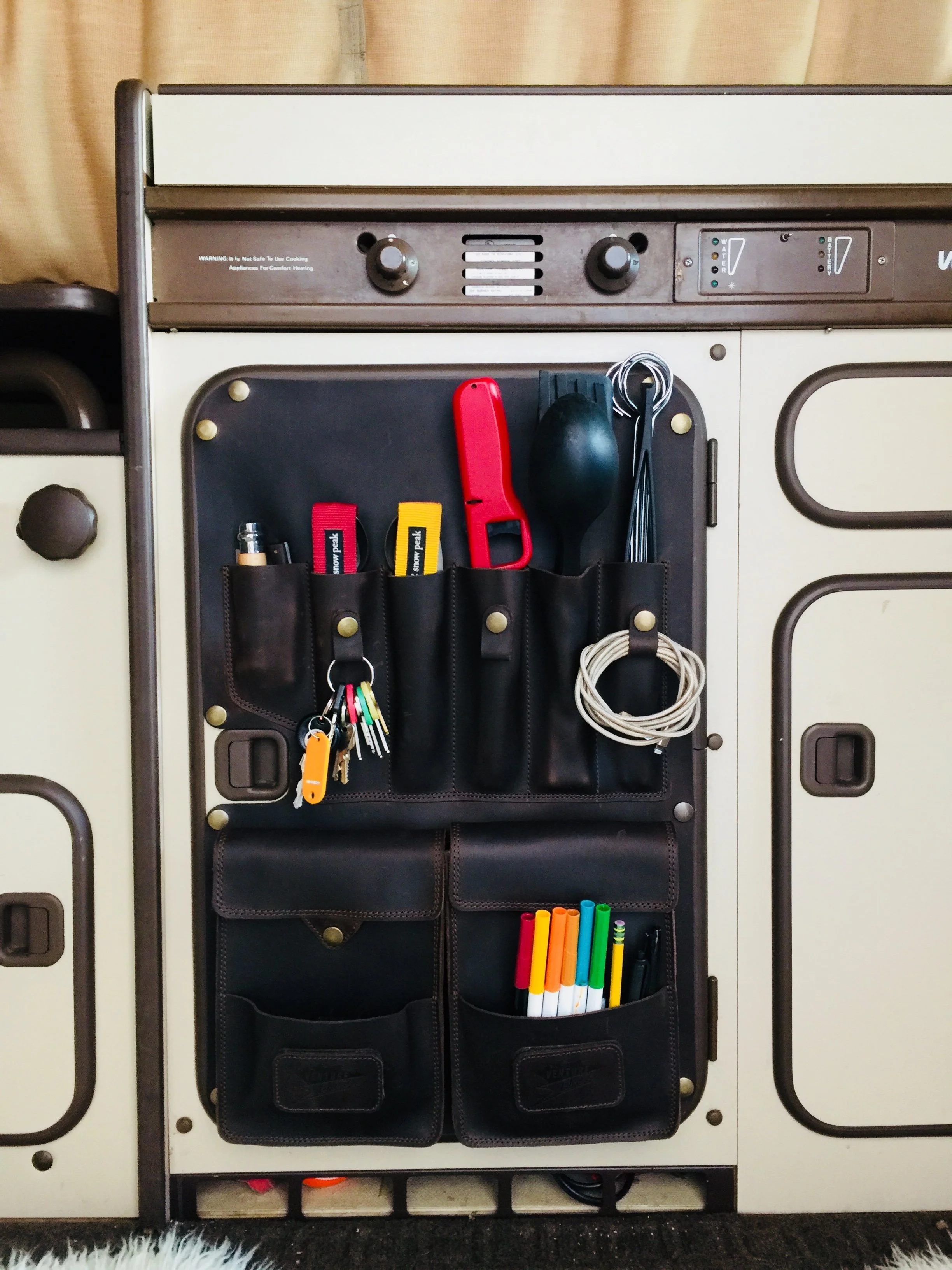 A storage unit attached to a vintage kitchen cabinet with various kitchen tools and supplies organized in pockets, including a scissors, pens, markers, wires, and a spatula.