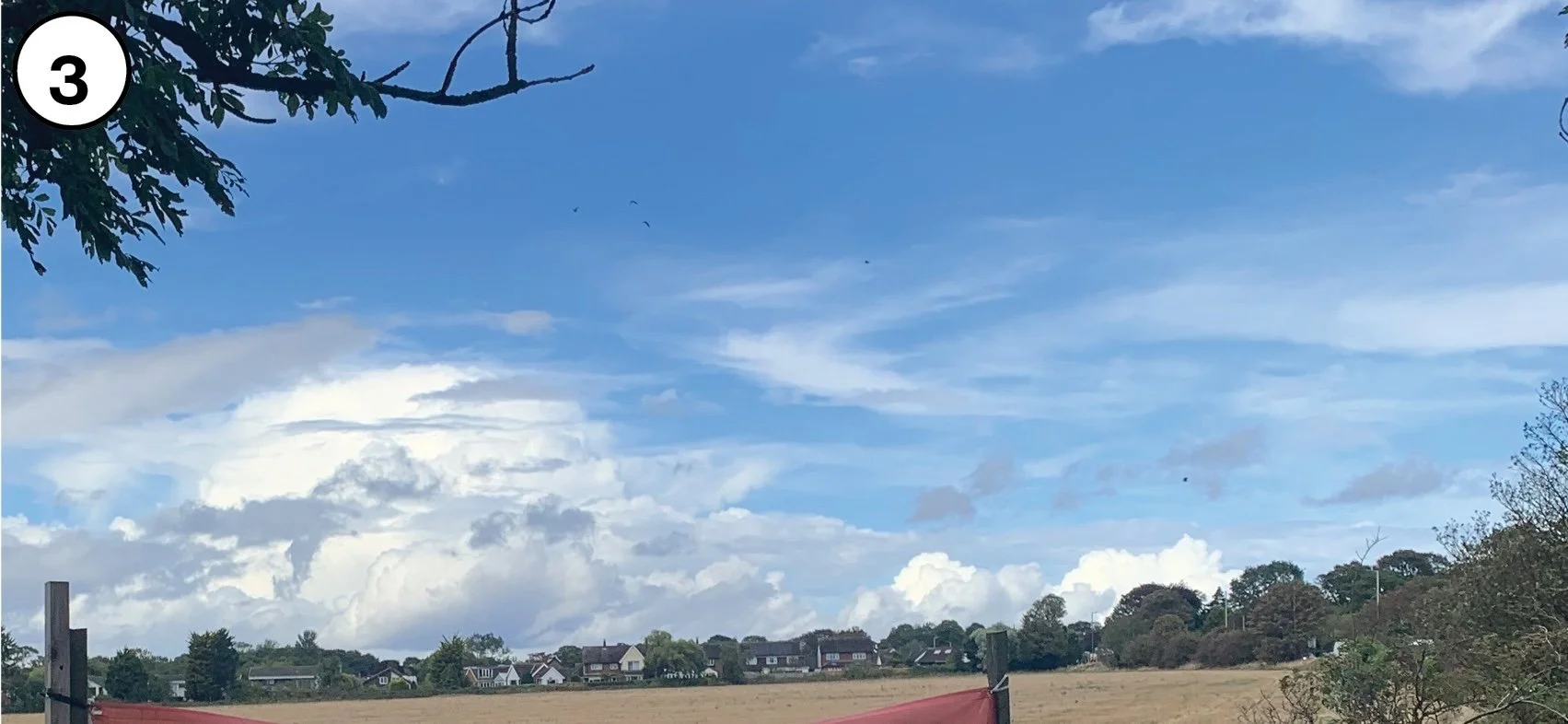 A wide outdoor view of a rural area with a clear blue sky, scattered clouds, and distant houses surrounded by greenery and trees.