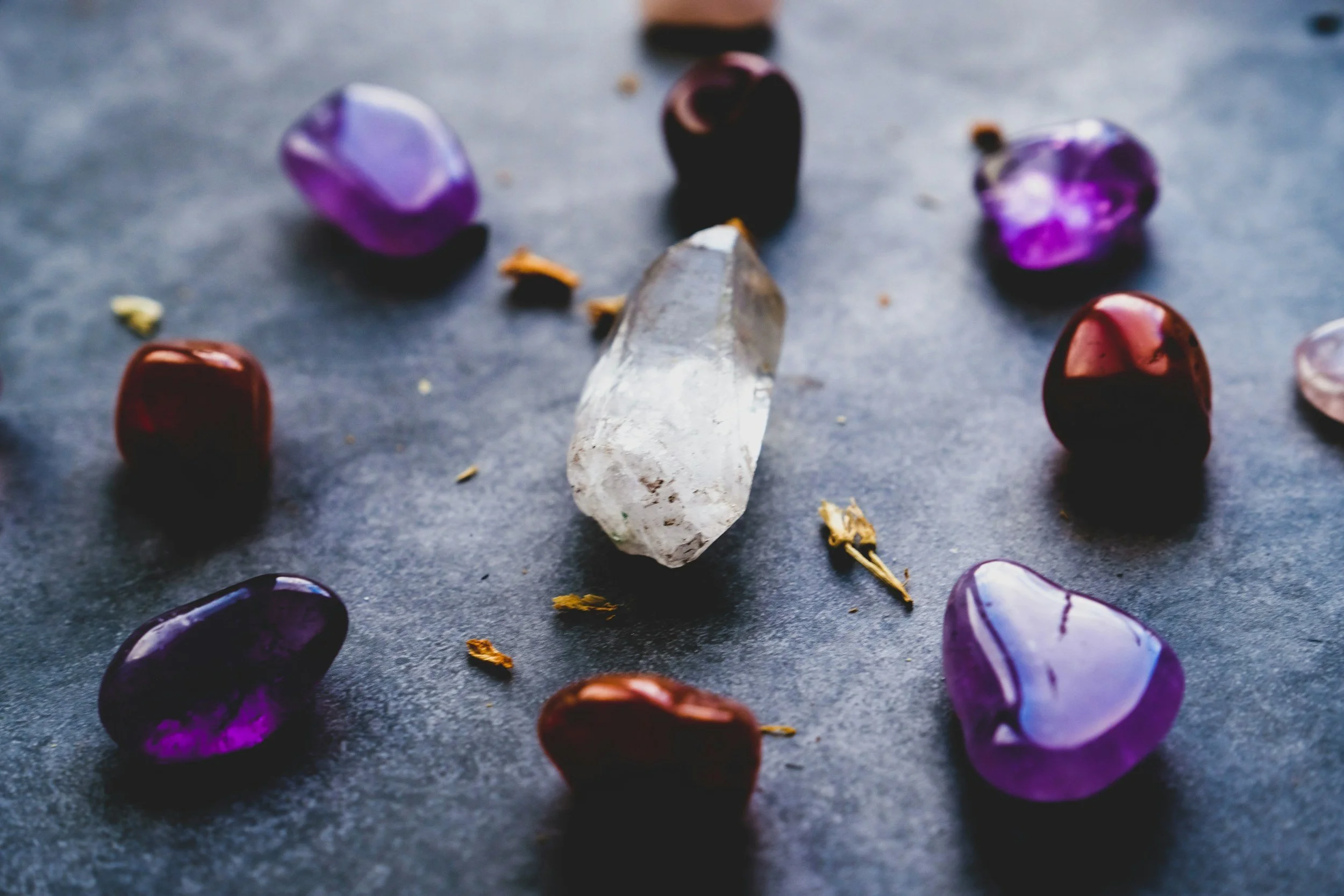 A clear quartz crystal surrounded by several purple and red tumbled stones on a gray surface.