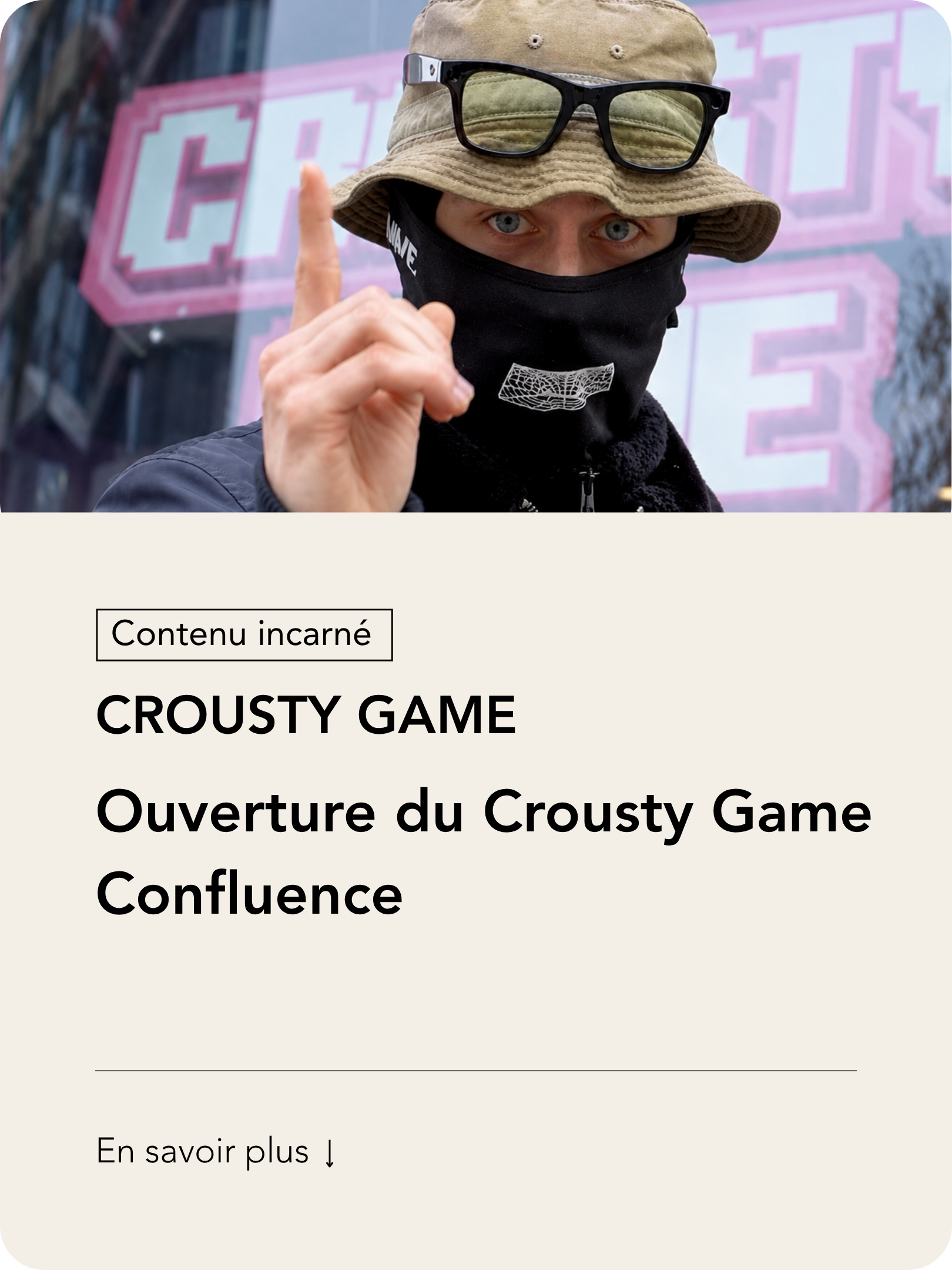 Un homme porte un chapeau beige, des lunettes noires sur la chapeau, un masque noir, et pointe du doigt en regardant l'objectif, devant une façade avec des lettres roses