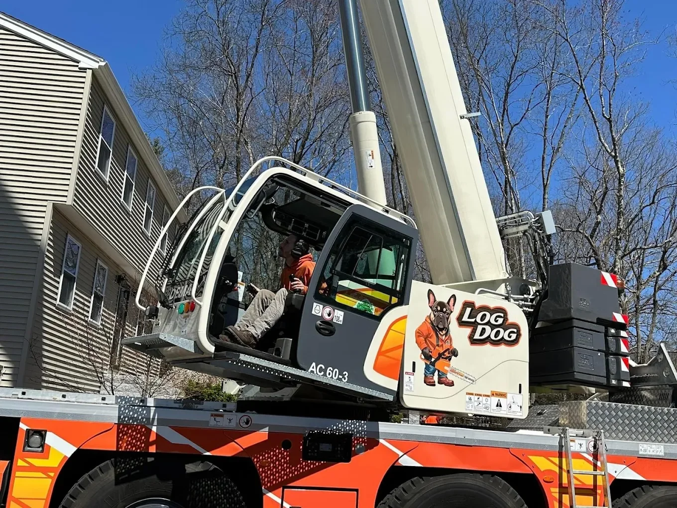Child sitting inside a large crane truck labeled 'Log Dog,' with a cartoon dog figure on the side, next to a residential house with trees in the background.