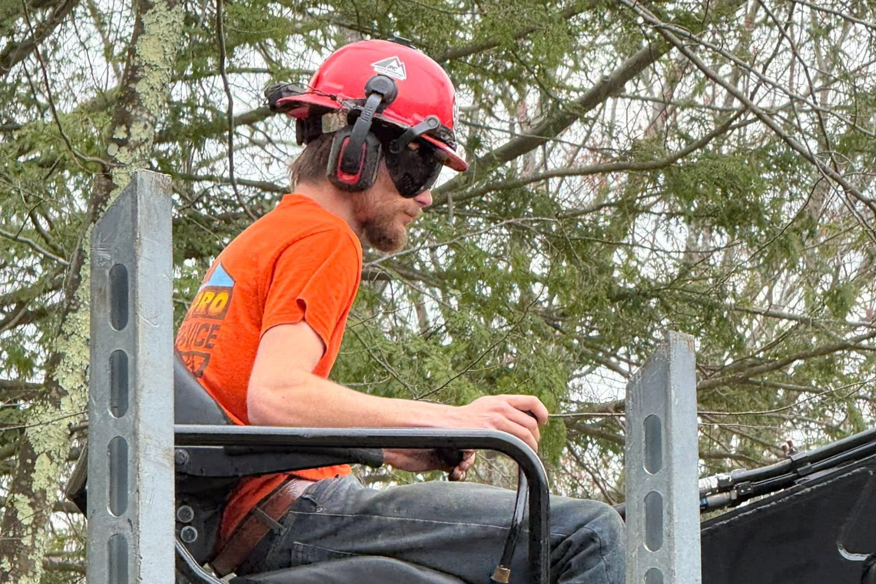 A man wearing a red safety helmet, black sunglasses, and an orange shirt is sitting on a platform, working with equipment outdoors in front of trees.
