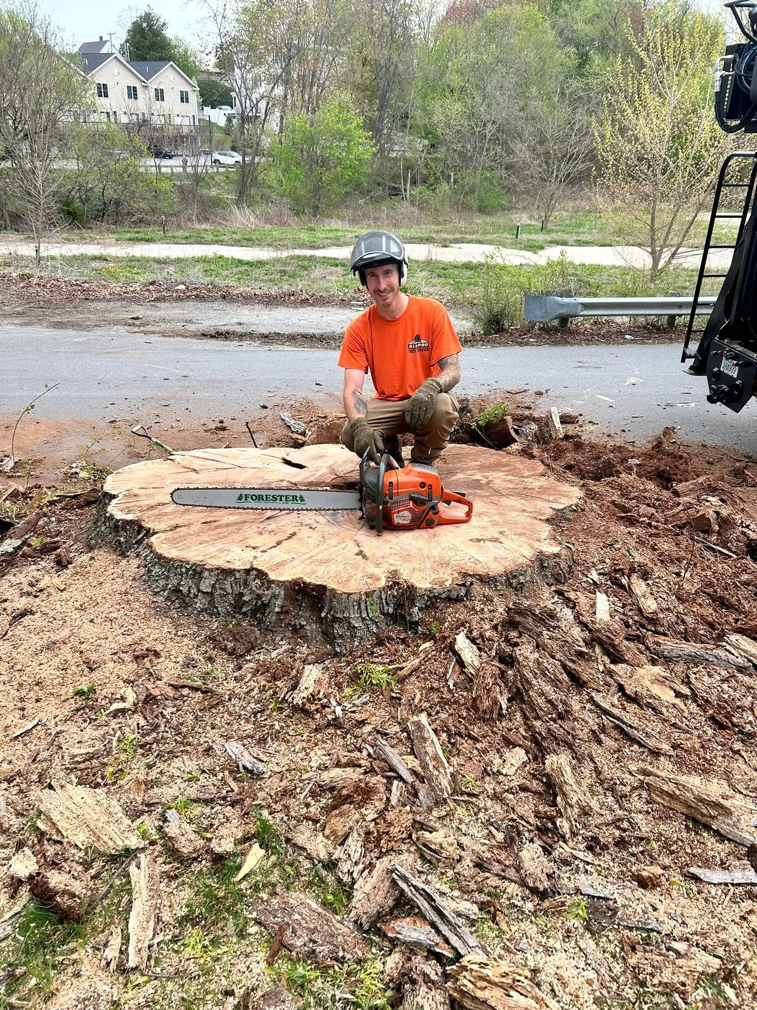 A man in an orange shirt and a helmet crouching next to a large cut tree trunk with a chainsaw resting on the trunk, in an outdoor setting with trees and buildings in the background.