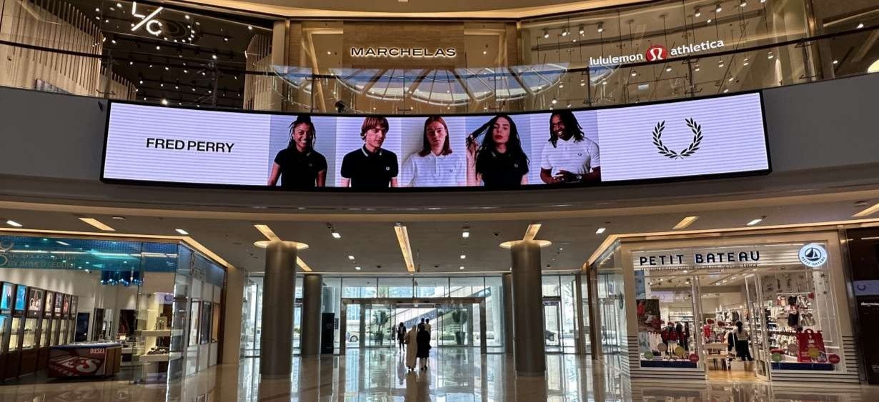 Interior of a shopping mall with a large digital screen displaying five people, including Fred Perry branding, and stores like Petit Bateau visible.