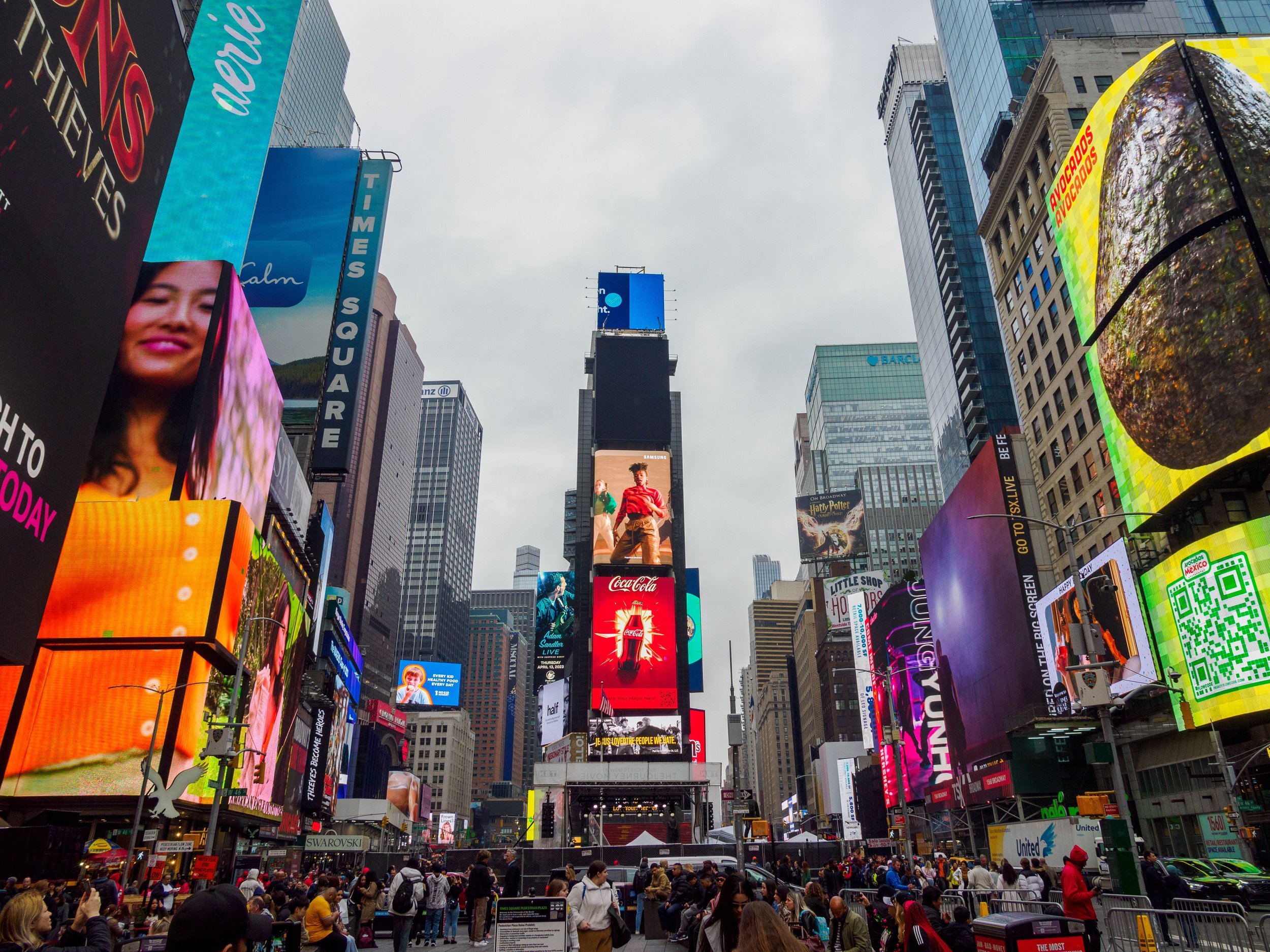 Crowds of people in Times Square, New York City, surrounded by large digital billboards and tall buildings, on an overcast day.