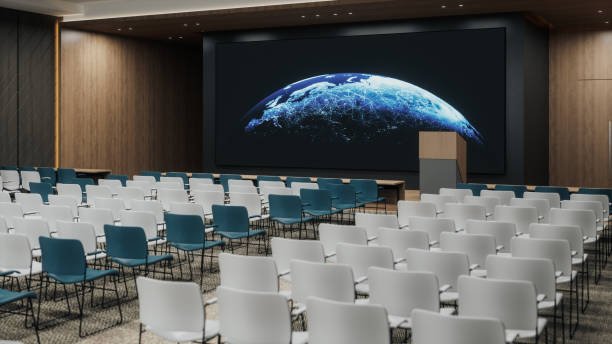 Empty conference room with a large screen displaying the Earth from space, rows of white and blue chairs facing the screen.