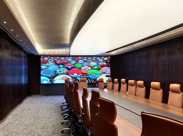 A conference room with a long wooden table and tan leather chairs, dark wood-paneled walls, and a large screen displaying colorful test tubes or capsules.