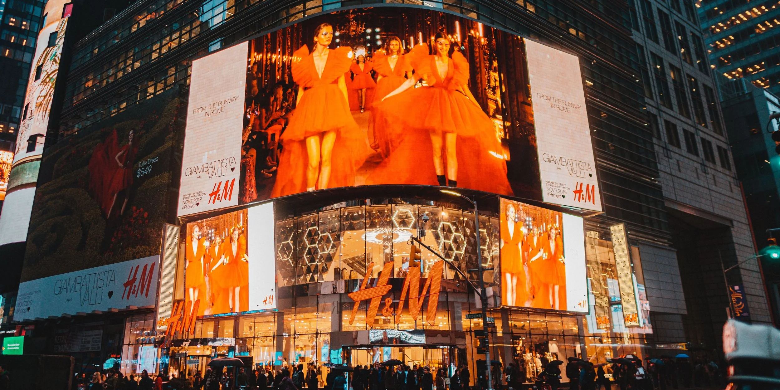Digital billboard in Times Square displaying models walking in an orange dress fashion show. Crowds of people with umbrellas are below, and surrounding buildings are visible.
