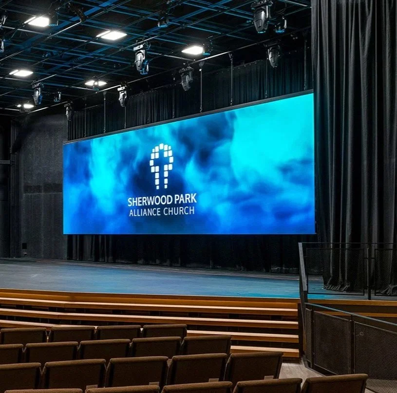 Interior of a church auditorium with a large digital screen displaying the logo and name of Sherwood Park Alliance Church, surrounded by black curtains and rows of empty brown seats.