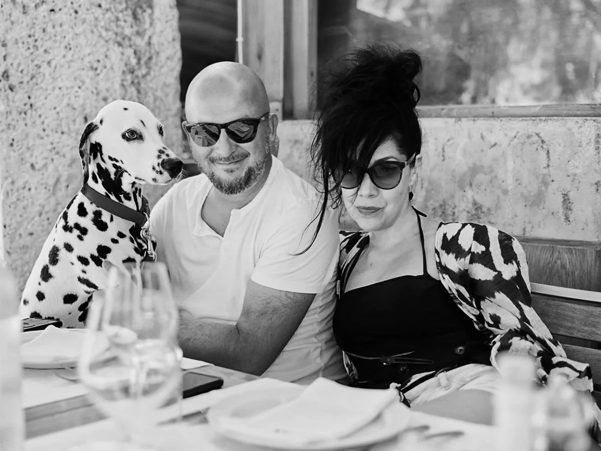 A black-and-white photo of a man, woman, and Dalmatian dog sitting at a restaurant table. The man is bald, wearing sunglasses and a white t-shirt, smiling. The woman has dark, wavy hair in a voluminous updo, wearing large sunglasses and a patterned top, looking into the camera. The Dalmatian sits beside the man, looking away. There are wine glasses, plates, and a phone on the table.