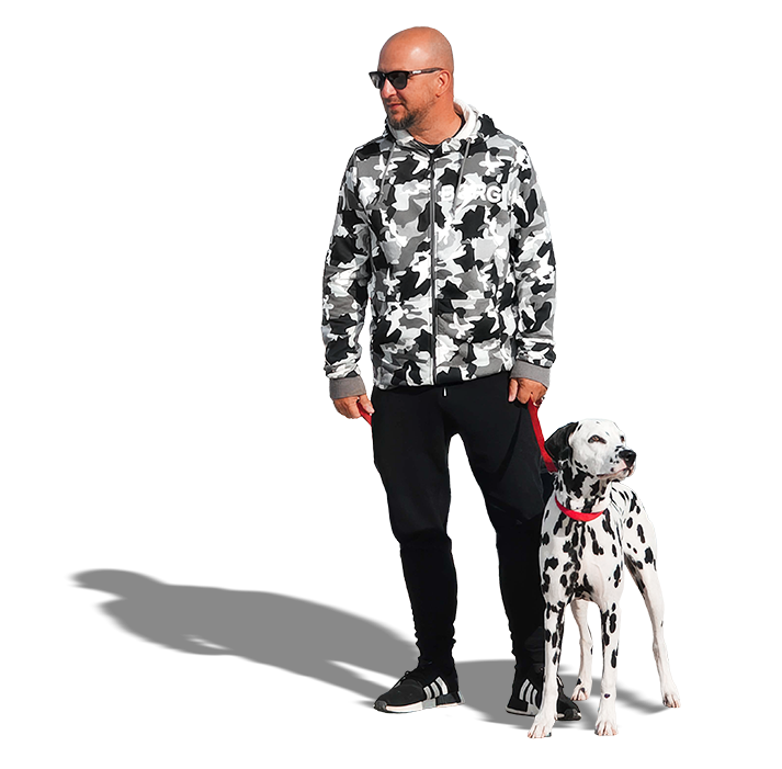 A man with sunglasses wearing a camouflage hoodie and black pants standing next to a Dalmatian dog on a red leash.