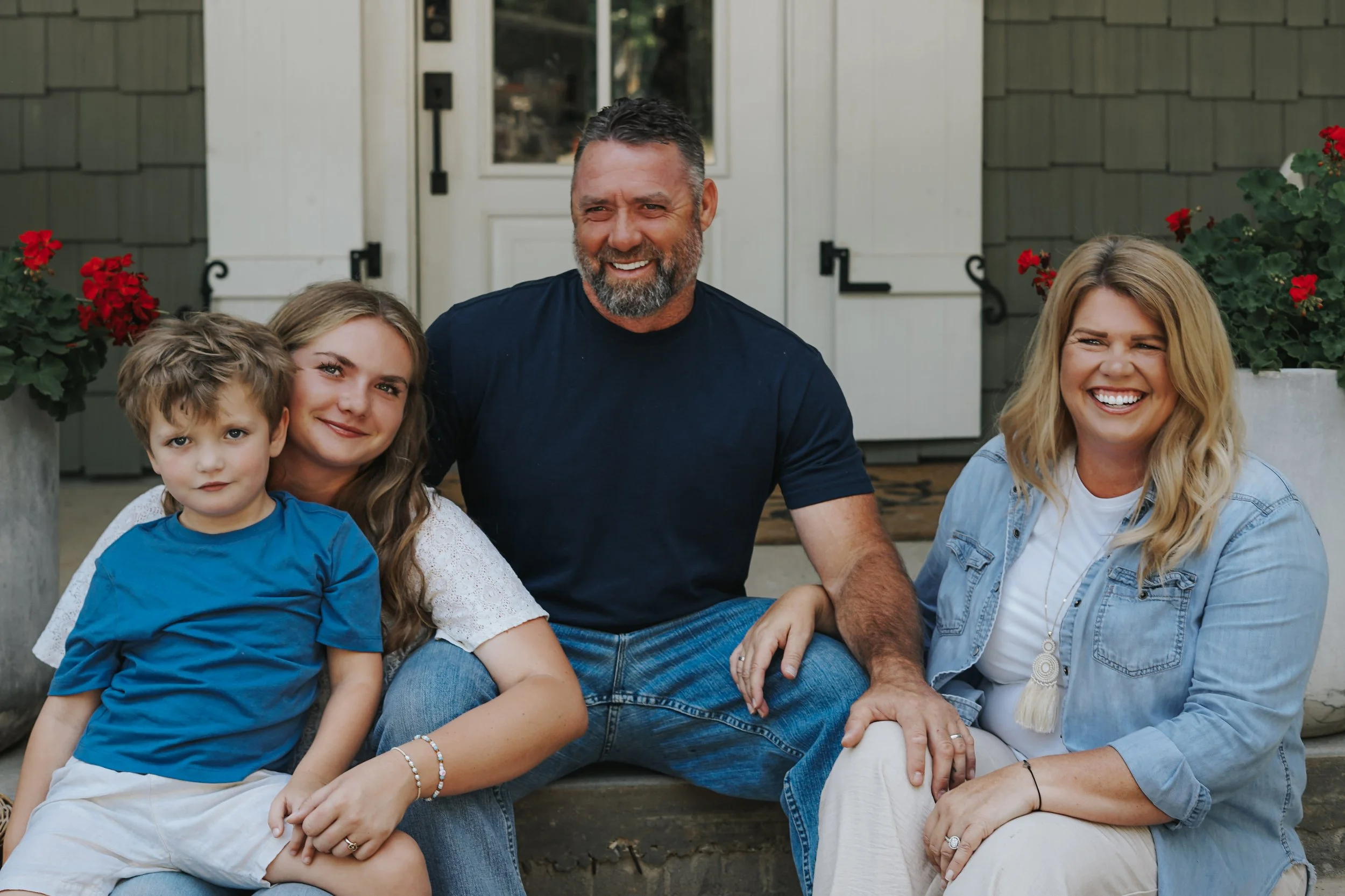 A smiling family of five sitting on steps outside their house, with a man, woman, two girls, and a boy, surrounded by red flowers in planters.