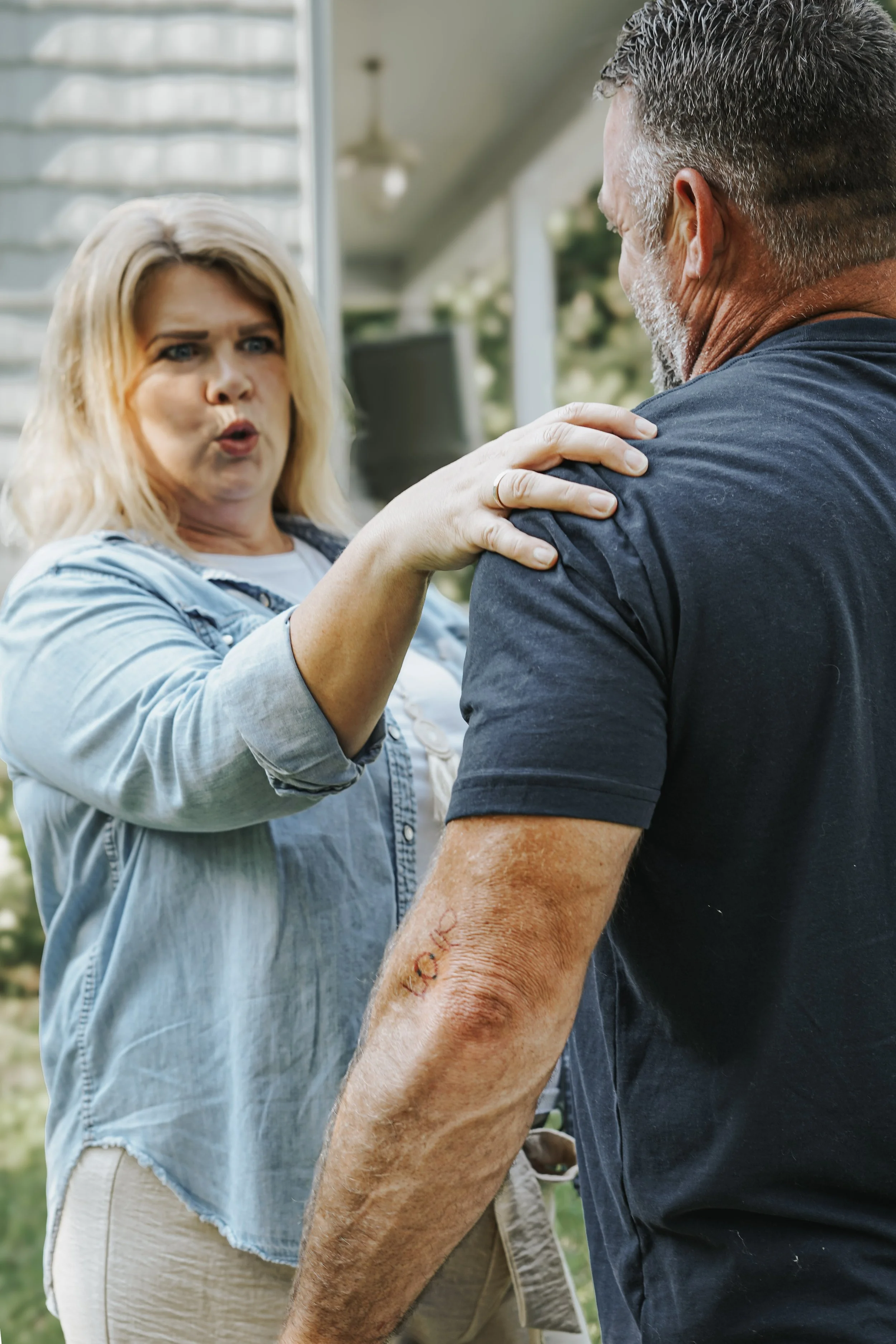 A woman with blonde hair and a man with gray hair are engaged in an emotional conversation outdoors. The woman has her hand on the man's shoulder, and the man has a fresh scar on his arm.