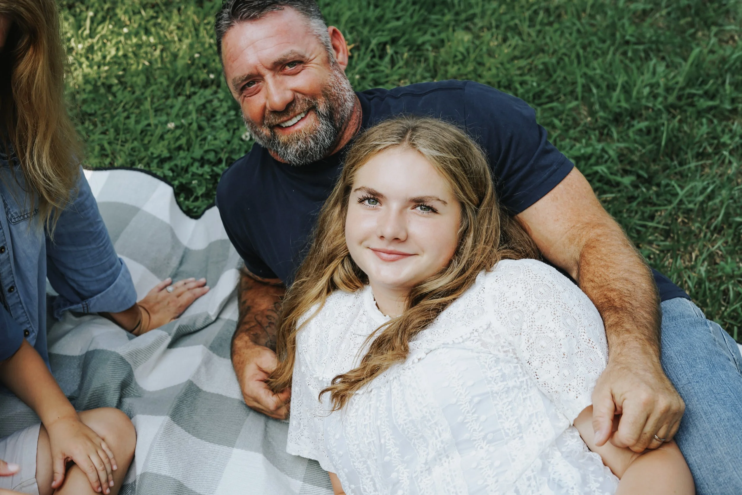 A man with gray hair and beard lying on his side on a blanket, smiling, with a young girl with long blonde hair, blue eyes, and a white dress lying on her back next to him, both outdoors on green grass.
