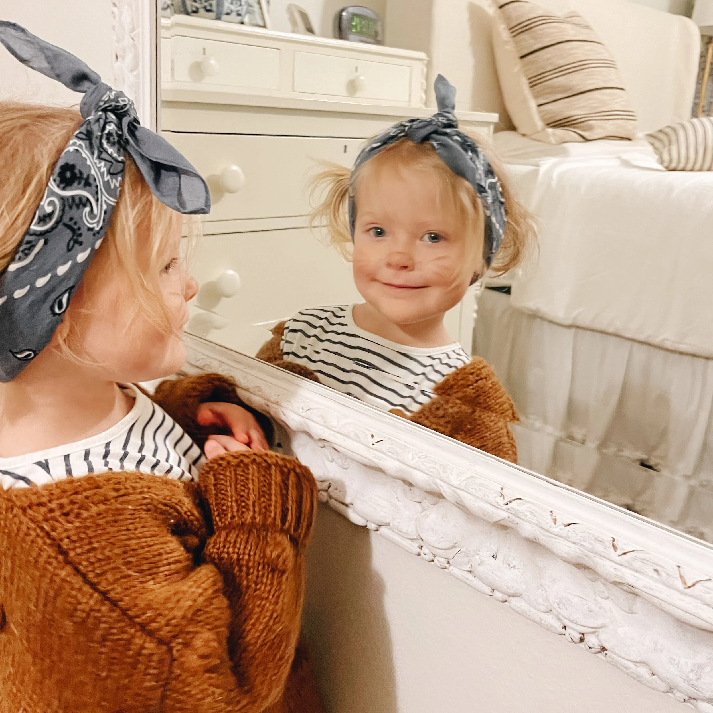 A young girl with curly blonde hair, wearing a striped shirt and a gray headscarf, looking at a mirror in a bedroom, with part of a brown teddy bear visible in the foreground. The background includes a white dresser with a small green digital clock and a bed with striped pillows.