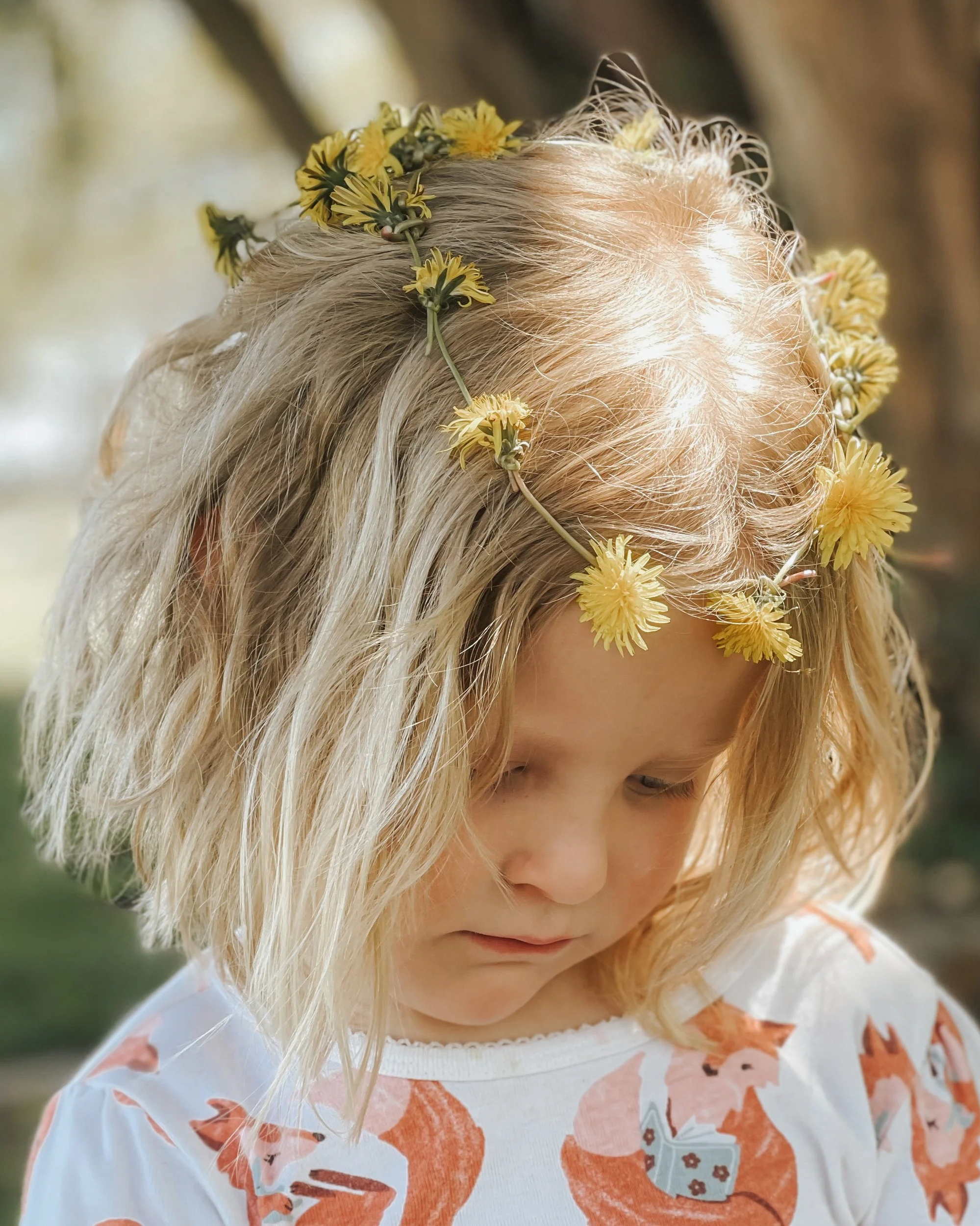A young girl with blonde hair wearing a flower crown made of yellow blossoms, looking downward, outdoors.