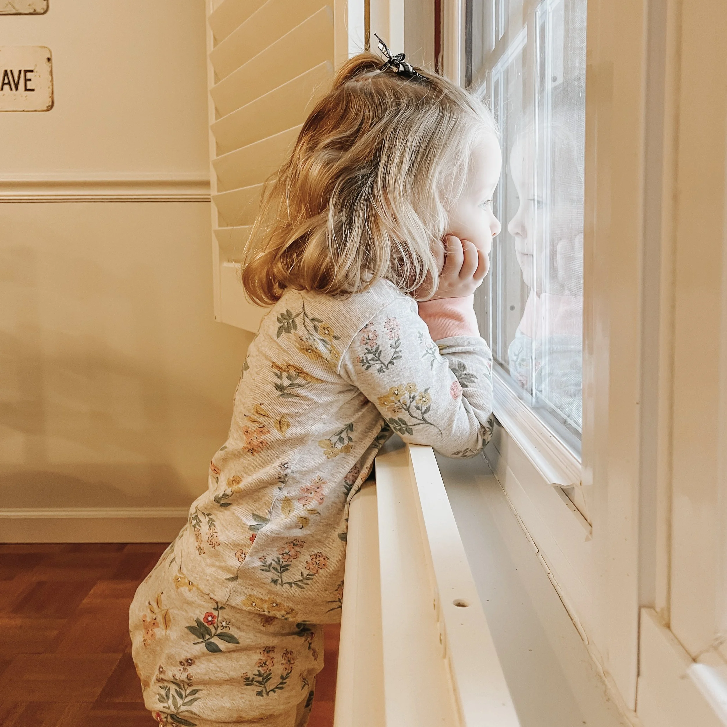 A young girl with curly blonde hair gazes out of a window with horizontal blinds, resting her chin on her hands.