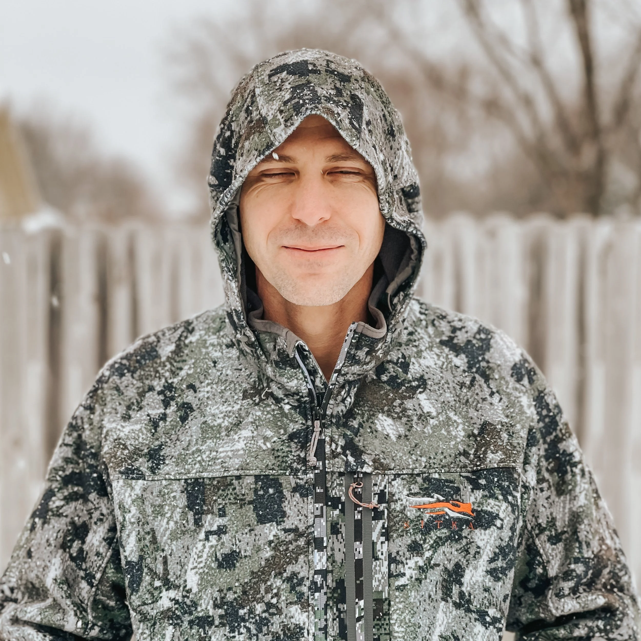A smiling man wearing a camouflage-patterned jacket and a camouflage hat on a snowy day with blurred trees and a fence in the background.