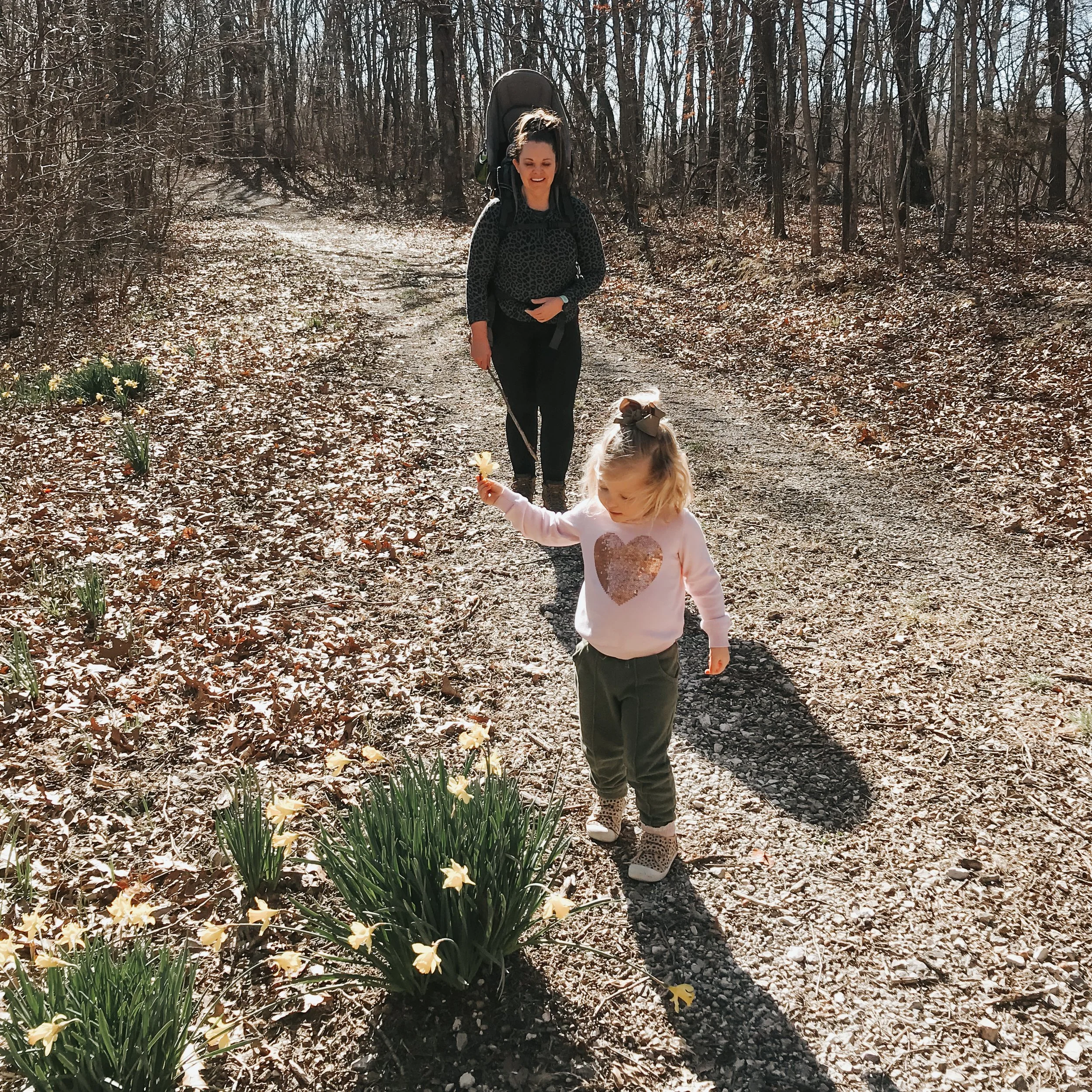 A woman with dark hair tied back, wearing a black leopard print top and black pants, walks on a forest trail with a child holding her hand, another child with blonde hair and pink jacket walking nearby, in a wooded area during daytime with bare trees and yellow flowers