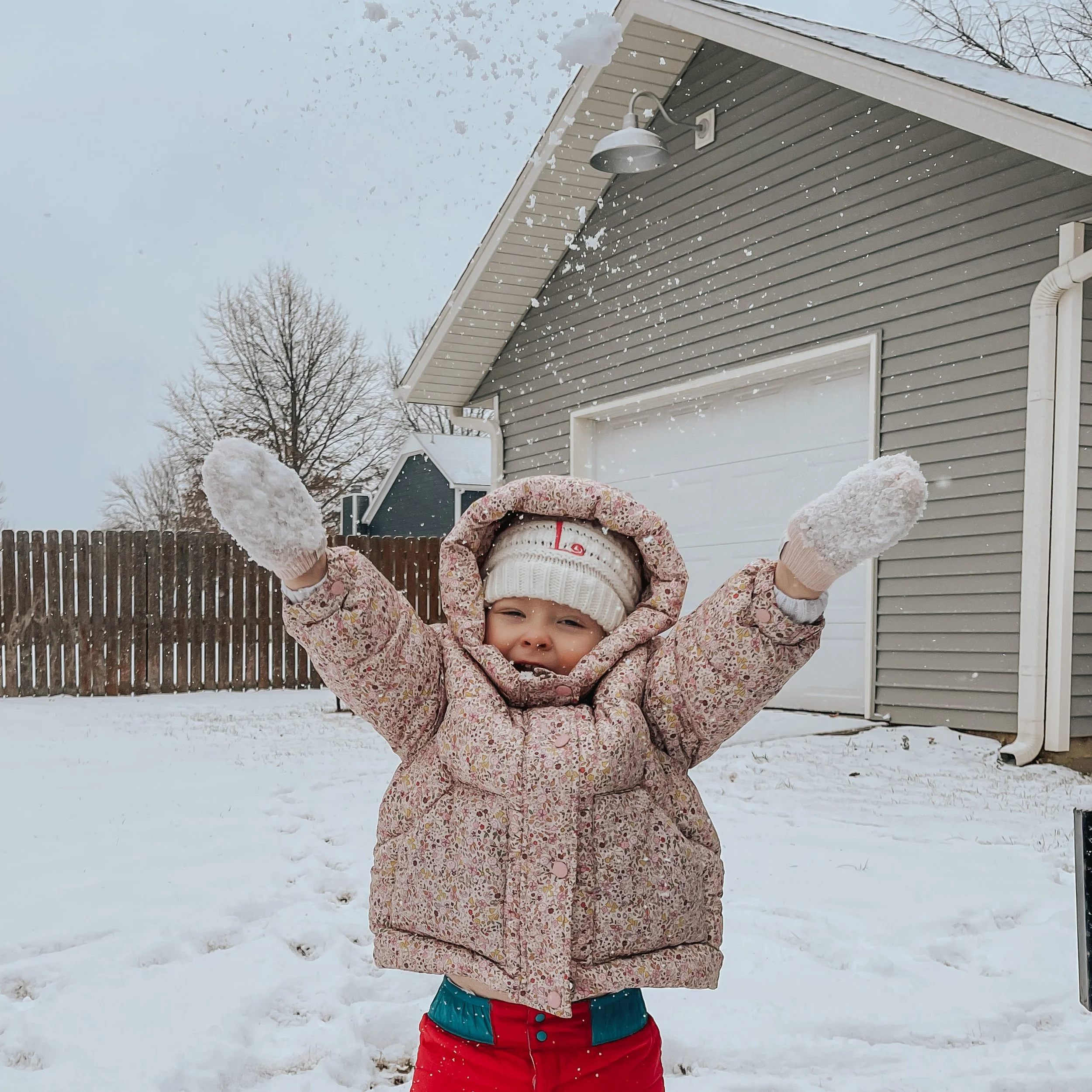 A young girl wearing a warm jacket, beanie, and mittens smiling and holding snow in the outdoors during snowy weather.