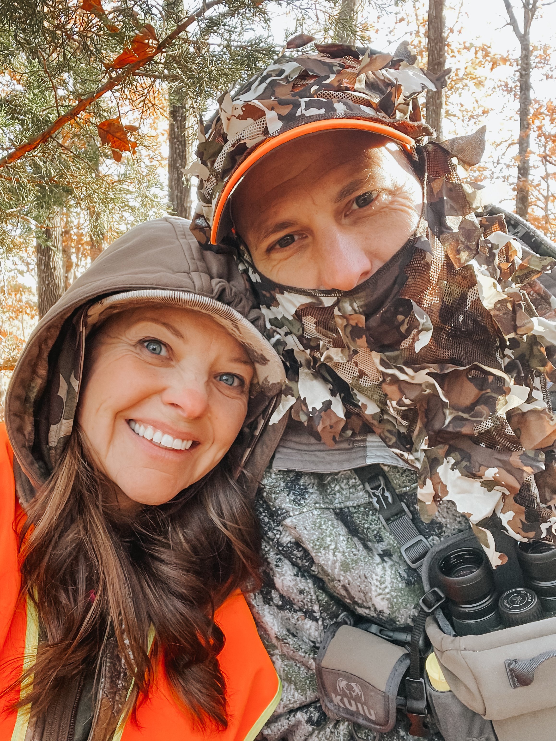 Two women hiking in a forest, smiling and posing for a selfie. They are dressed in outdoor and camouflage gear, with trees and sunlight in the background.