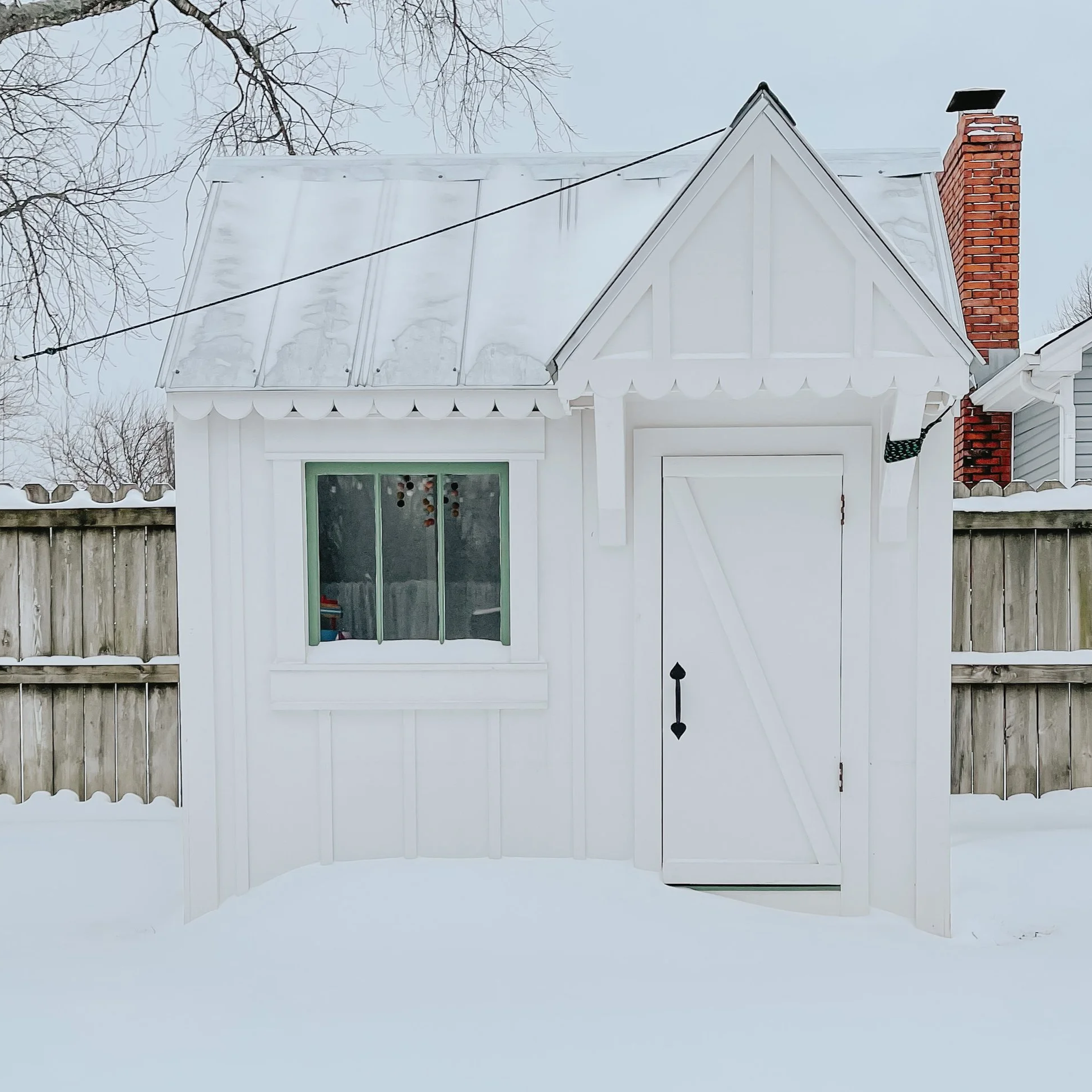 A white house with a gabled roof covered in snow, a brick chimney, a window with a green frame, and decorative horizontal siding, with tree branches and a grey sky in the background.