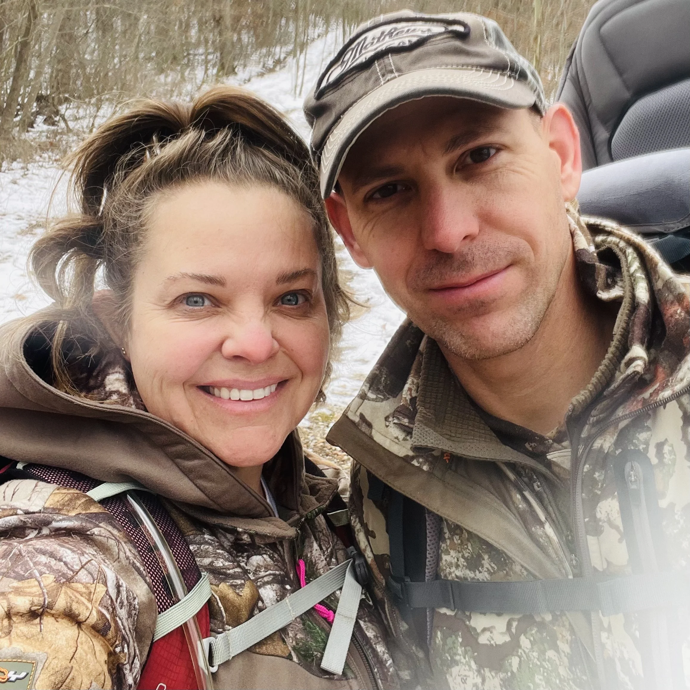 Close-up of a smiling woman and a man smiling outdoors, both wearing camouflage clothing, surrounded by trees with snow on the ground.