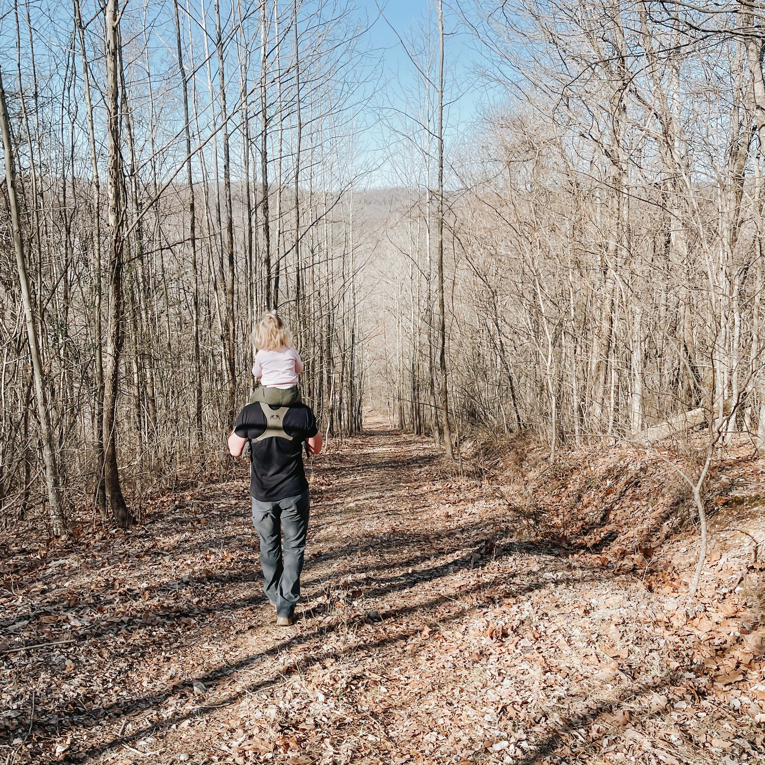 A person with curly blonde hair walking along a trail in a leaf-covered forest during winter, with leafless trees and a blue sky in the background.