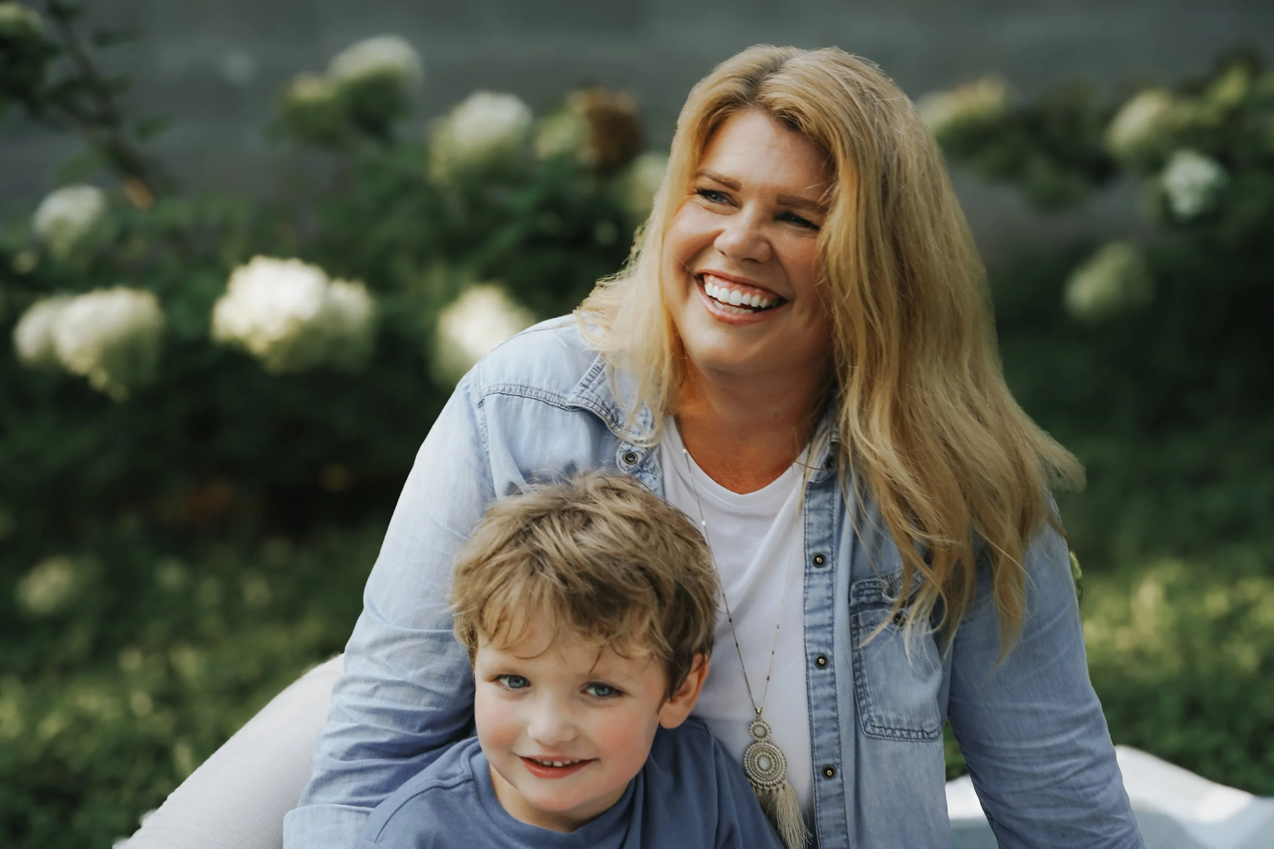 A woman with blonde hair smiling outdoors with a young boy in front of her, surrounded by greenery and white flowers.