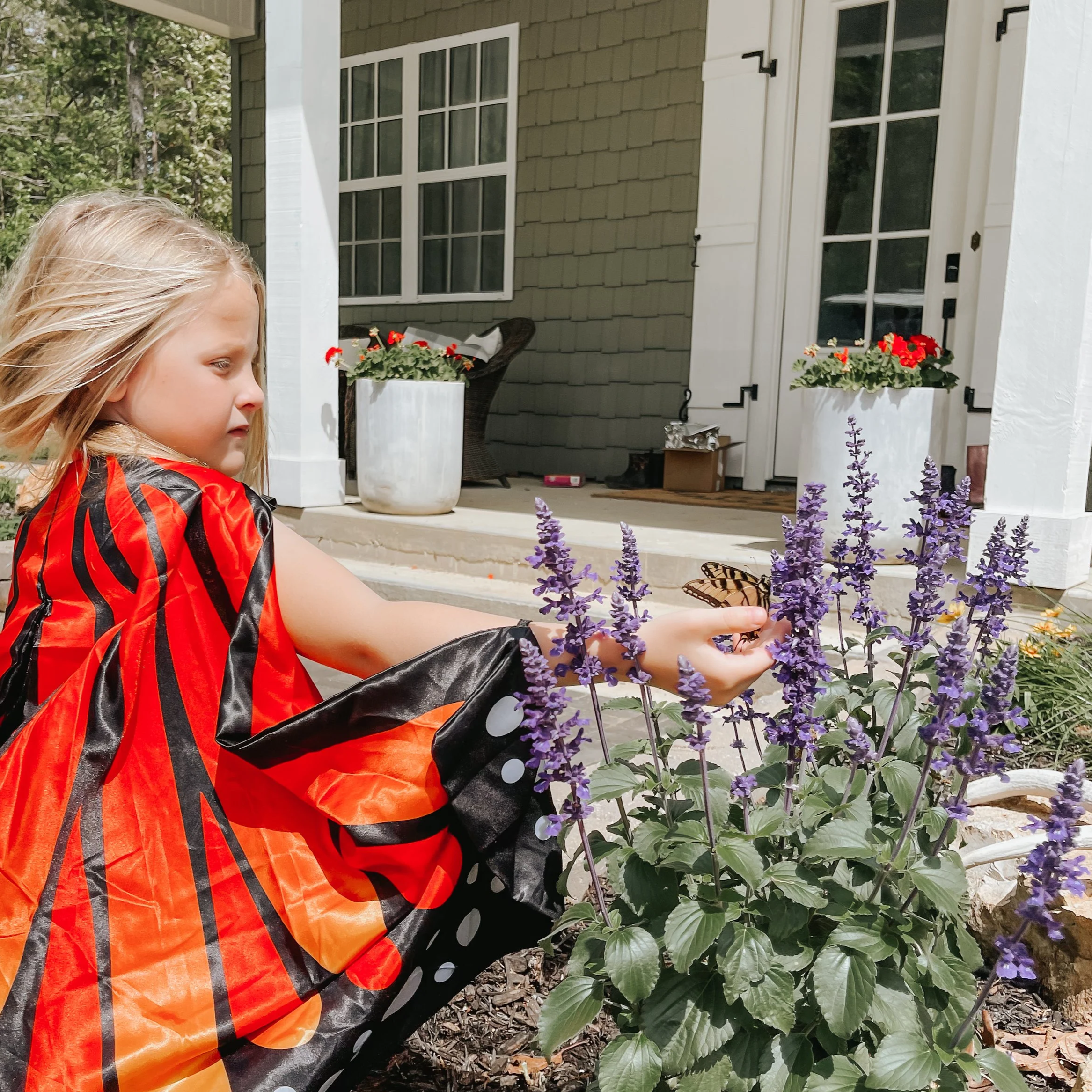 A young girl with blonde hair and a red and black shirt is outside near a flower bed with purple flowers, a white planter with red flowers, and trees in the background.