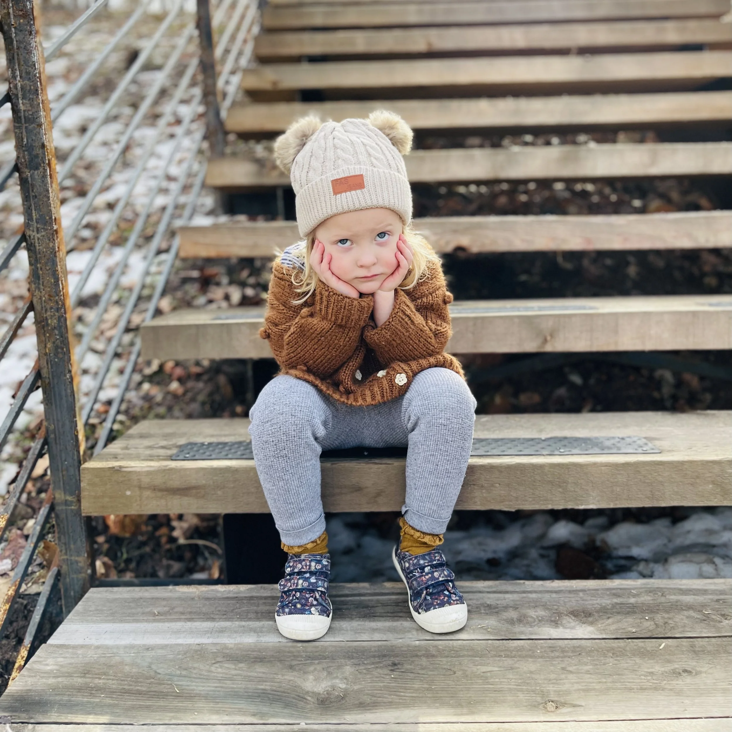 Young girl with blond hair wearing a beige knit hat with pom-poms, sitting on outdoor wooden stairs with hands on her cheeks, looking up with a thoughtful expression.