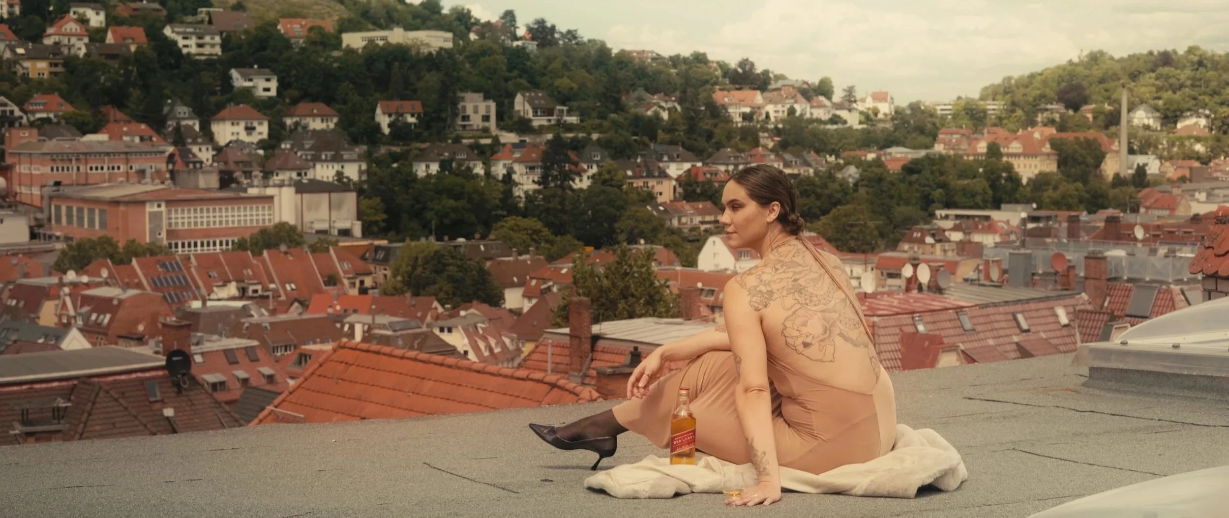 A woman with tattoos and dressed in a beige dress, sitting on a rooftop with a cityscape in the background. She has black heels and a drink bottle beside her, with a blanket underneath.