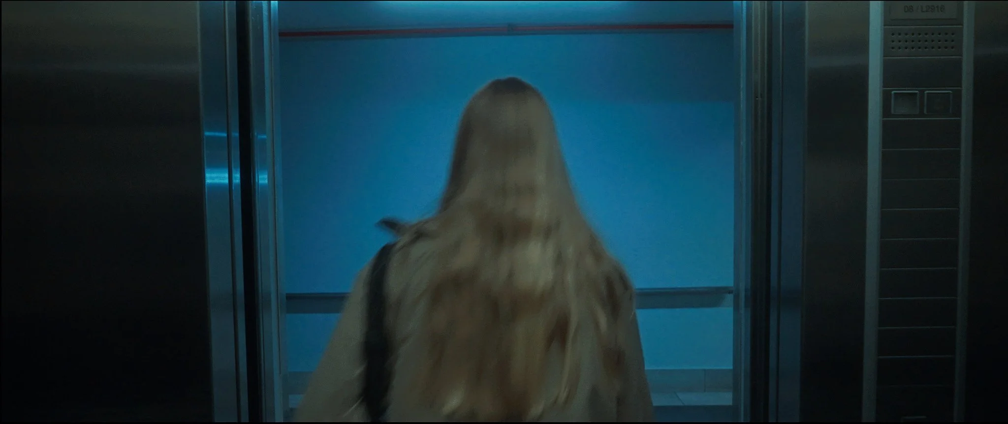 A woman with long blonde hair, carrying a shoulder bag, is entering an elevator.