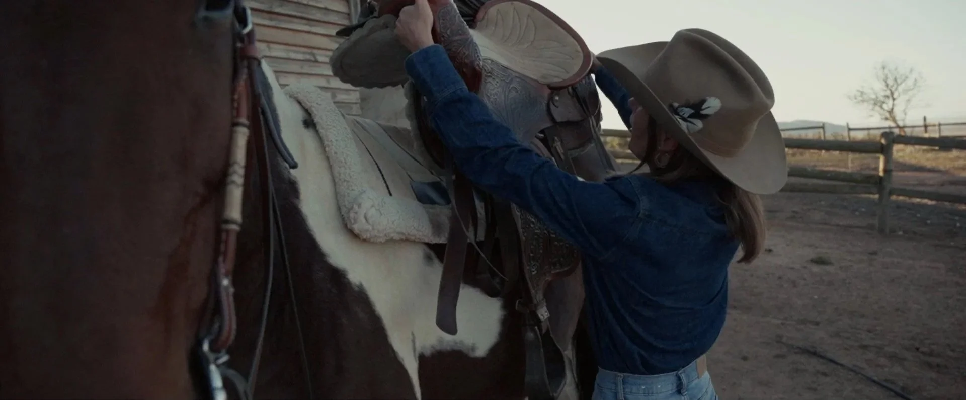 A person in a cowboy hat helping to saddle a horse on a ranch. The scene is set outdoors with a fence and a leafless tree in the background.