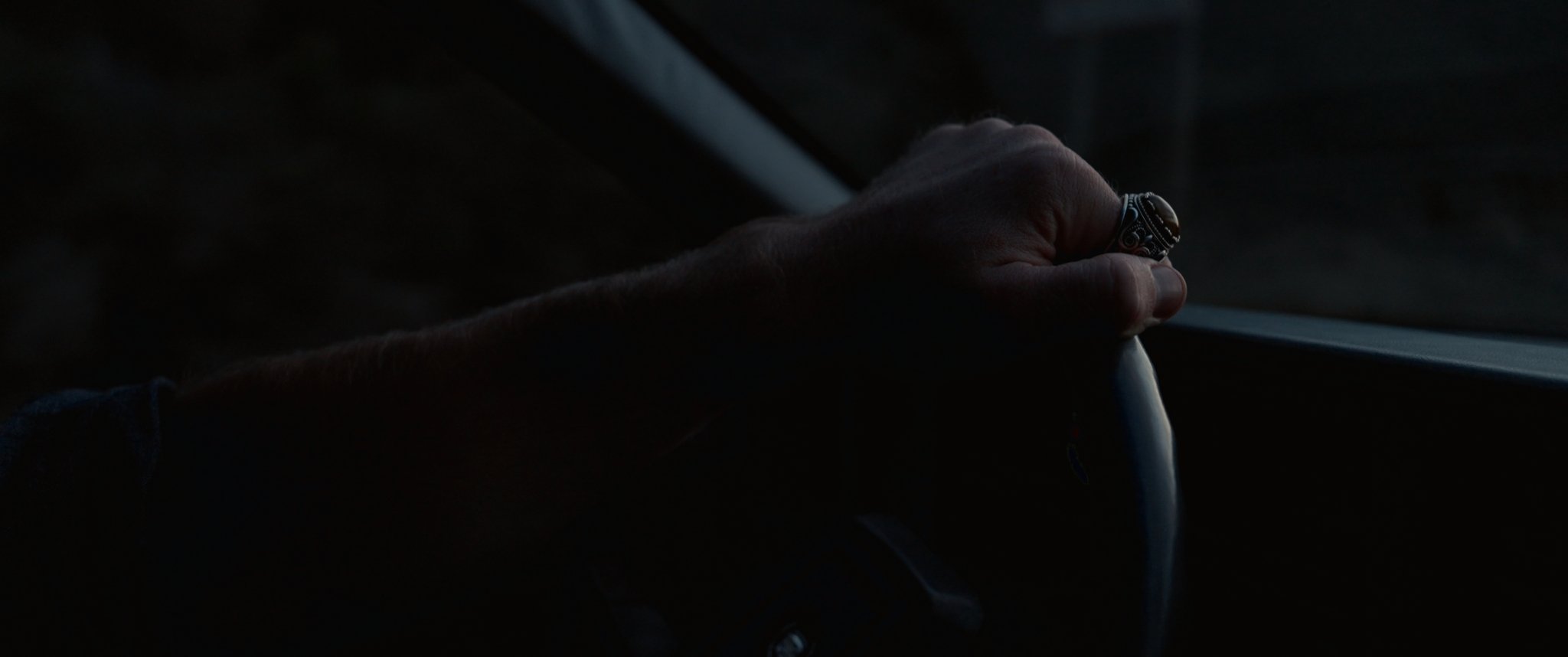 Close-up of a person's hand gripping a steering wheel in low light, wearing a silver ring with black details.