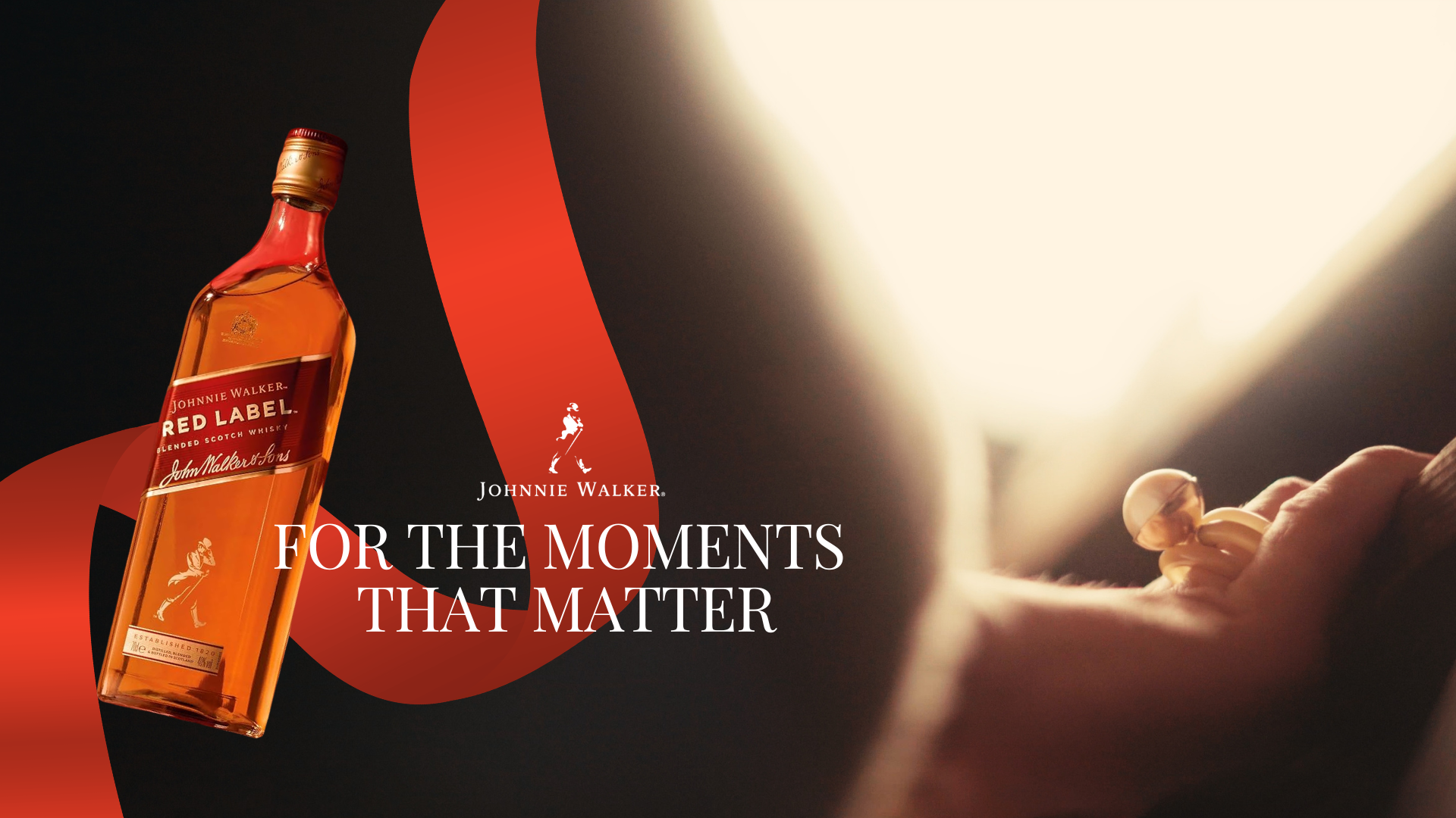 A bottle of Johnnie Walker Red Label blended Scotch whisky with a hand lighting a match near the bottle. The background features a dark setting with a bright light source illuminating part of the scene. The text reads 'For the moments that matter'.