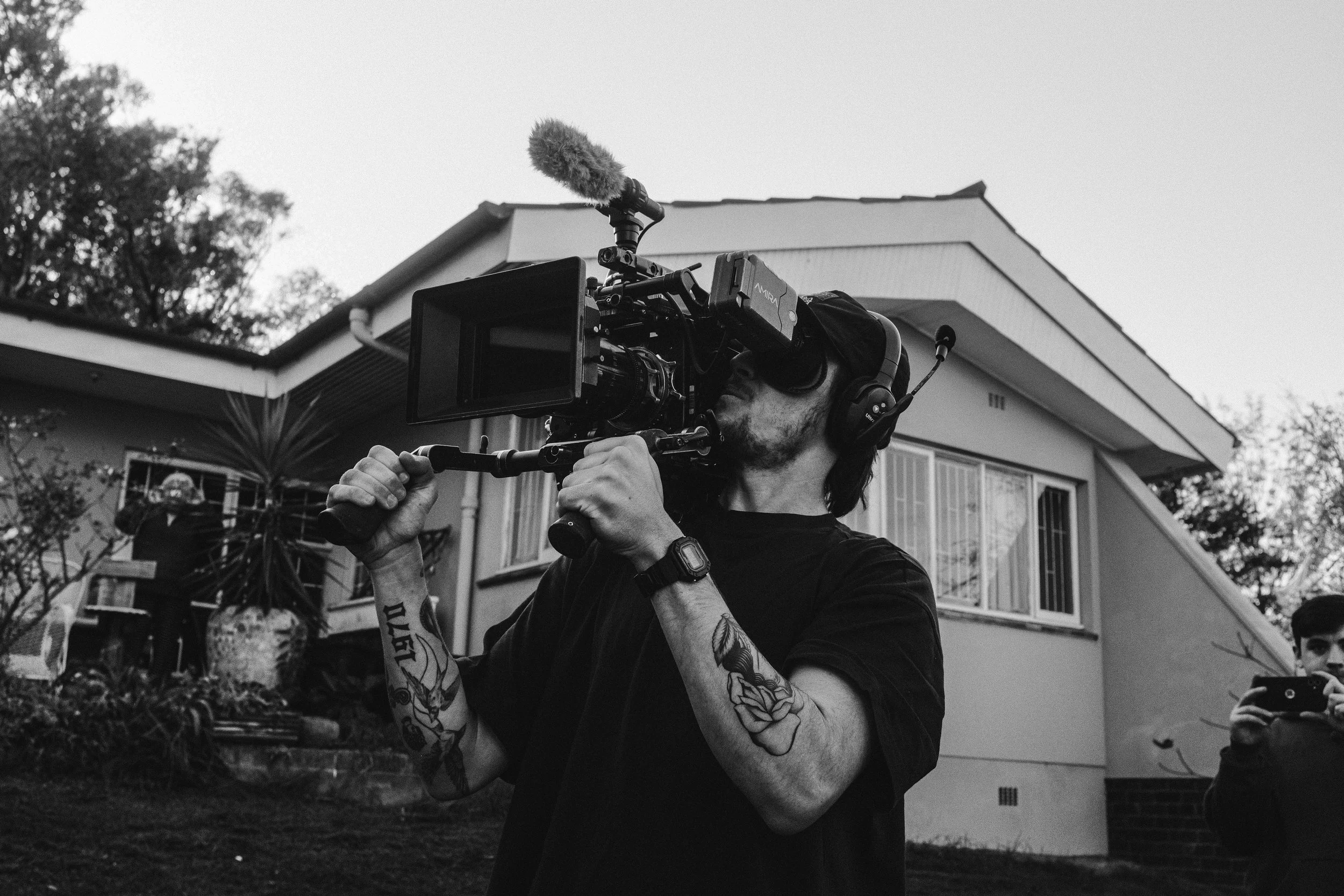 Ein Mann mit Tattoos auf den Armen, trägt eine schwarze T-Shirt, Kopfhörer und eine schwarze Armbanduhr. Er hält eine professionelle Kamera mit Mikrofon auf einem Stativ. Im Hintergrund ist ein Haus mit Balkon und Fenstern, sowie Bäume und Pflanzen.
