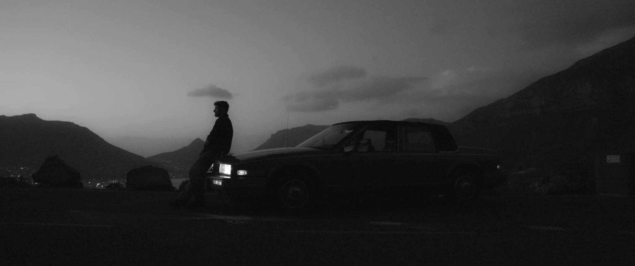 A black and white photograph of a man leaning against a car, standing in a mountainous landscape during dusk or dawn.