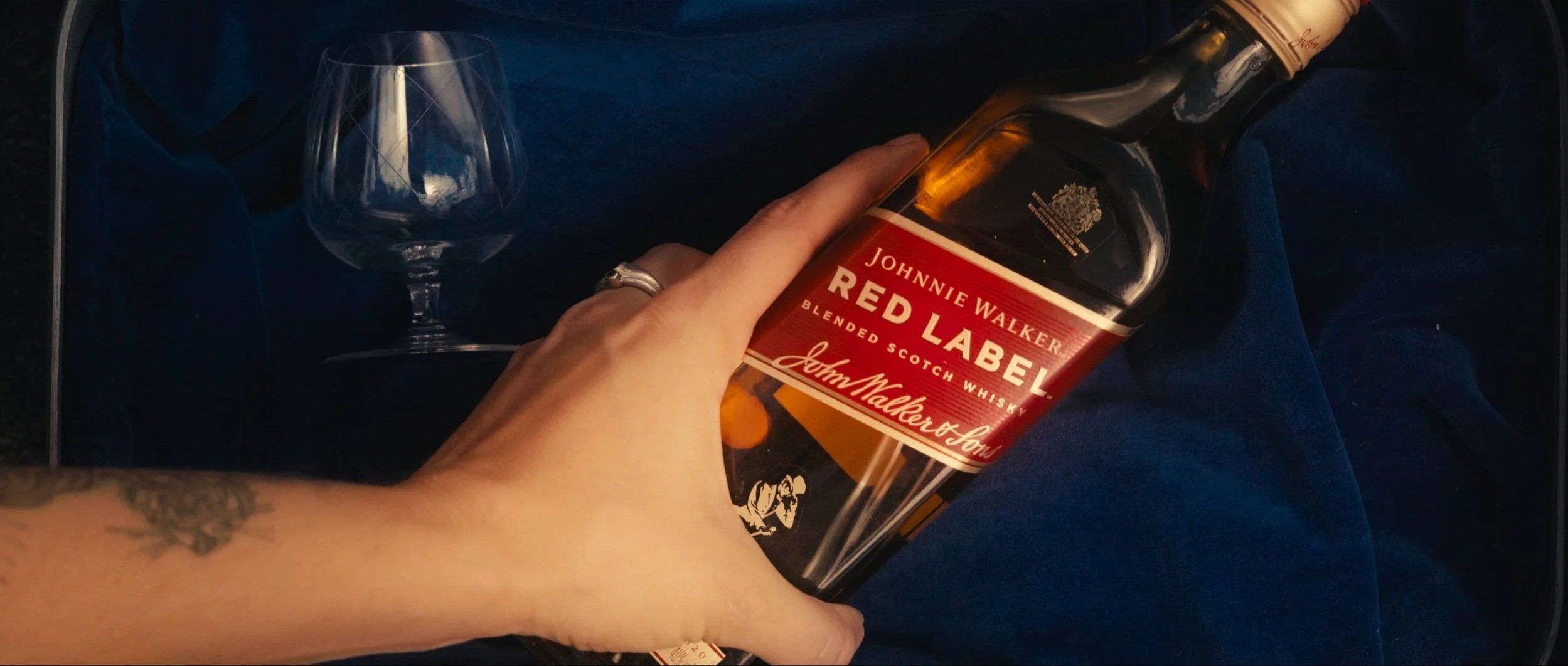 Person holding a bottle of Johnny Walker Red Label blended Scotch whisky, with a glass and blue fabric in the background.